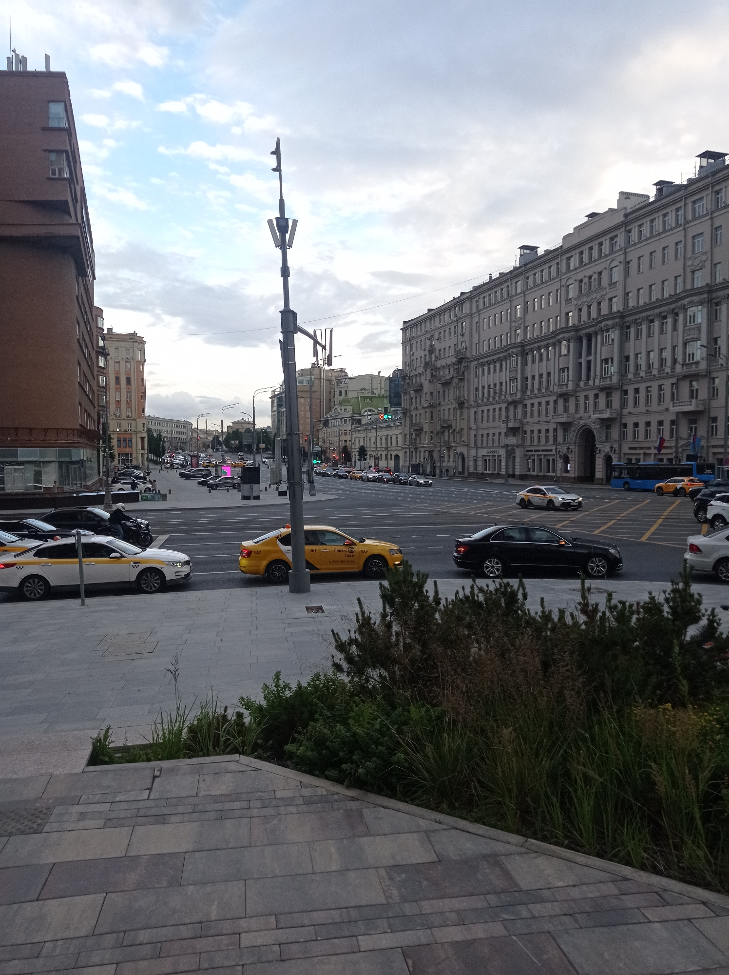 The image captures a street scene in Moscow, Russia, likely during the daytime under an overcast sky. In the foreground, a wide, grey paved area with geometric patterns transitions to a set of broad, grey stone steps. To the right, dense green bushes and tall grasses are visible. The middle ground features a busy intersection with multiple lanes of traffic. Several cars, including white and yellow taxis, are present on the road. A prominent tall, dark-colored traffic light pole stands to the right of the intersection. In the background, large, multi-story buildings line the streets. The building on the left is a reddish-brown, modern-looking structure. The building on the right is a grand, ornate, light-colored building with many windows and classical architectural details. The sky is partly cloudy with a muted grey and blue tone. The overall impression is one of a typical urban environment in Moscow, with significant traffic and imposing architecture.