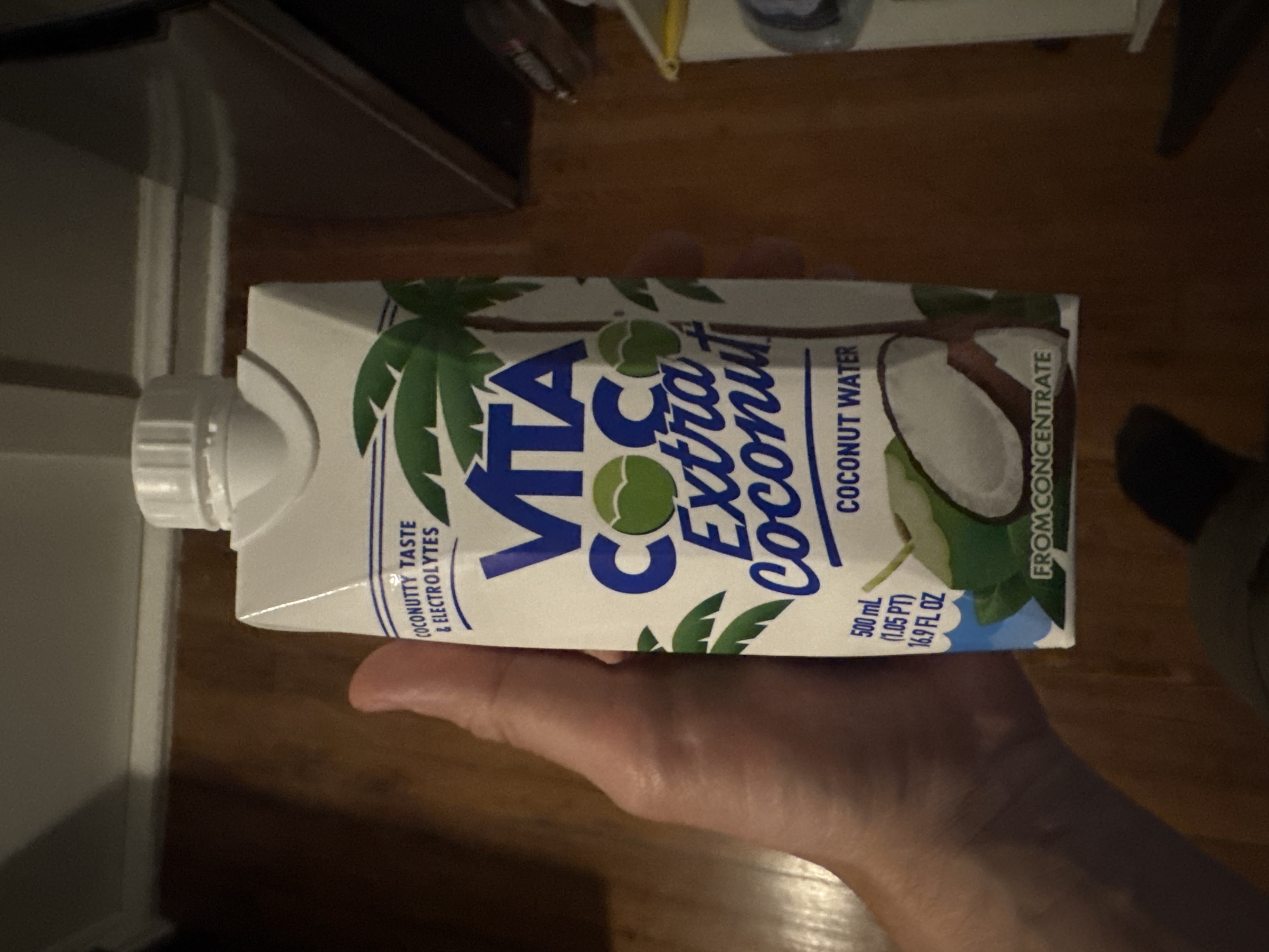 A hand is holding a carton of Vita Coco Extra Coconut water. The carton is white with blue and green lettering and images of palm trees and coconuts. The text on the carton reads "COCONUTTY TASTE & ELECTROLYTES", "VITA COCO Extra Coconut", "COCONUT WATER", "500 mL (1.05 PT) 16.9 FL OZ", and "FROM CONCENTRATE". The background appears to be a wooden floor and a wall with trim. The image is taken indoors. The location is not specified beyond Cleveland Heights, United States of America.