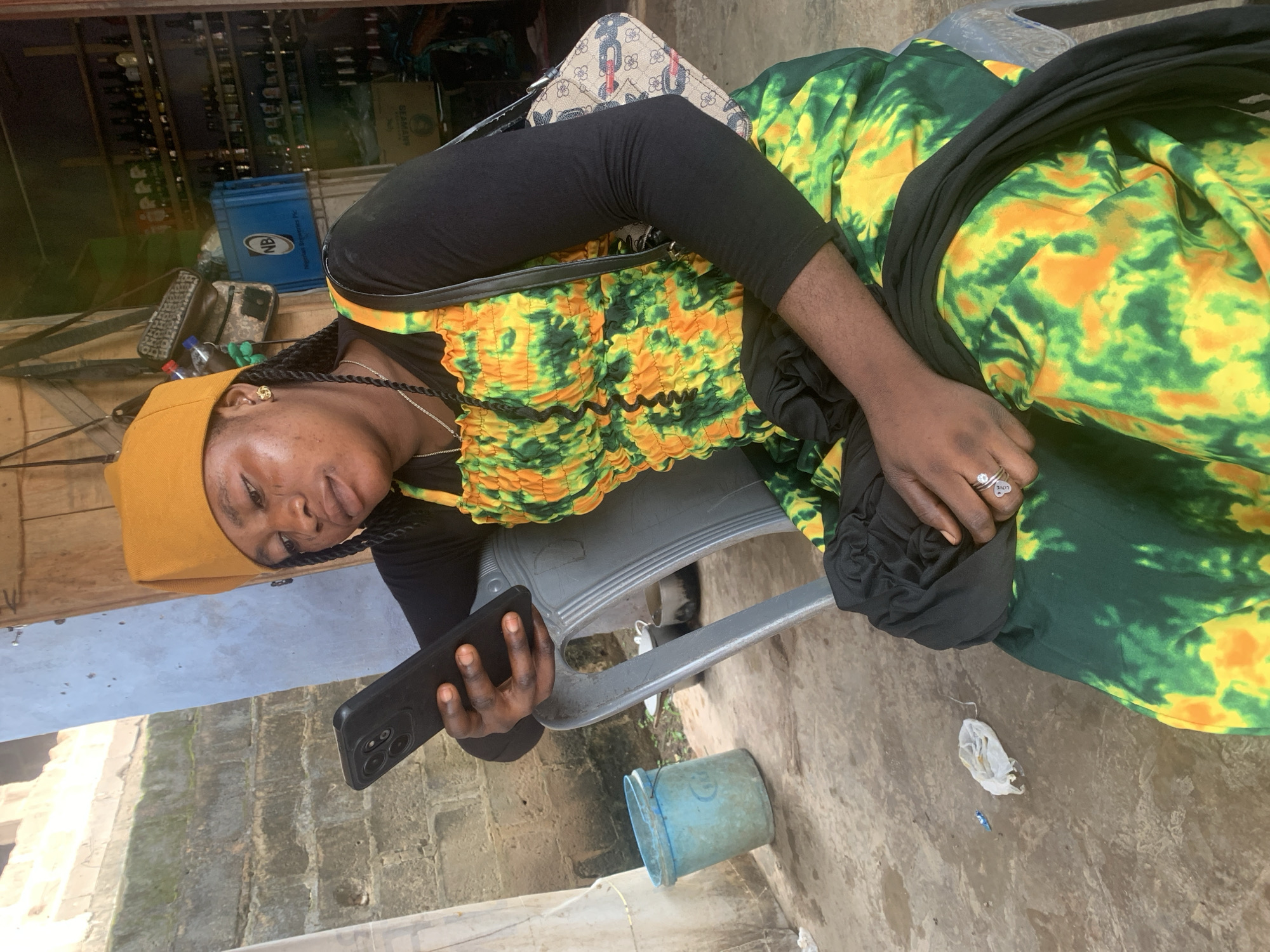 A young Black woman sits on a gray plastic chair, holding a black smartphone and looking at its screen. She wears a yellow cap, a black long-sleeved shirt, and a brightly colored dress with green, yellow, and orange tie-dye patterns. A dark fabric is draped over her lap. She has a ring on her left hand. To her left, on the ground, is a blue plastic bucket and some white debris. Behind her, shelves are stocked with bottles, suggesting a shop or bar. The overall environment appears to be an outdoor or semi-outdoor space in a town, potentially Shagamu, Nigeria, given the context.