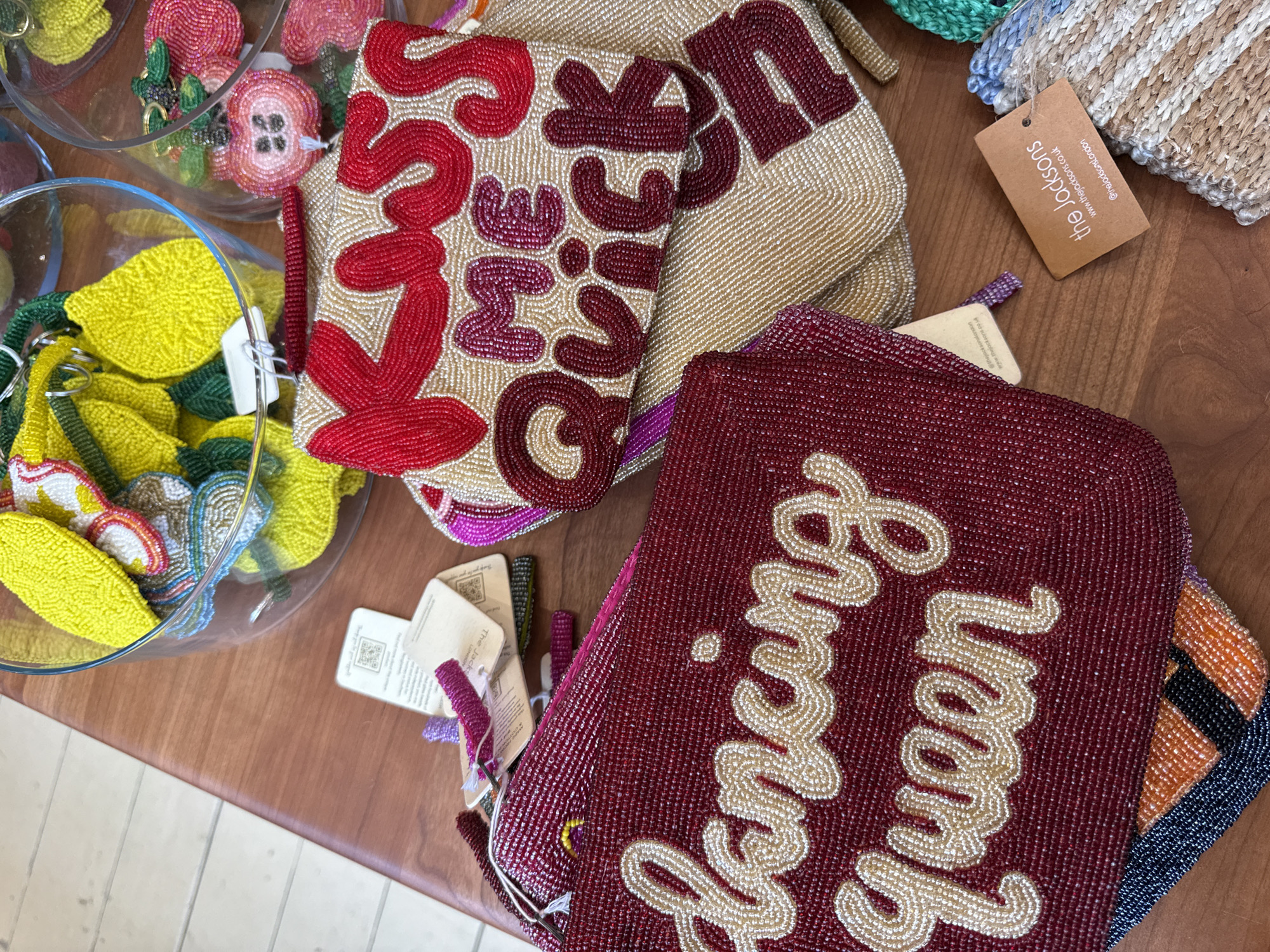 The image presents an overhead view of a retail display featuring various beaded accessories on a polished brown wooden surface. Two primary beaded clutch bags are visible: one deep red, embroidered with the text "Dancing Queen" in cream beads, and another beige clutch bag, embroidered with "KISS ME QUICK" in red and magenta beads. Additional beaded bags, including one with black, orange, and blue patterns, are partially visible beneath these. Two clear glass bowls, positioned in the upper right and upper left, contain multiple small, fruit-shaped beaded keychains, predominantly yellow lemons and pink-red apples. Several product tags are attached to the items, including a brown tag prominently displaying "the jacksons," with their website "www.thejacksons.co.uk" and Instagram handle "@thejacksonslondon," indicating a business presence in London, United Kingdom. A light-colored floor with visible seams is present in the background. No individuals are depicted in the scene.