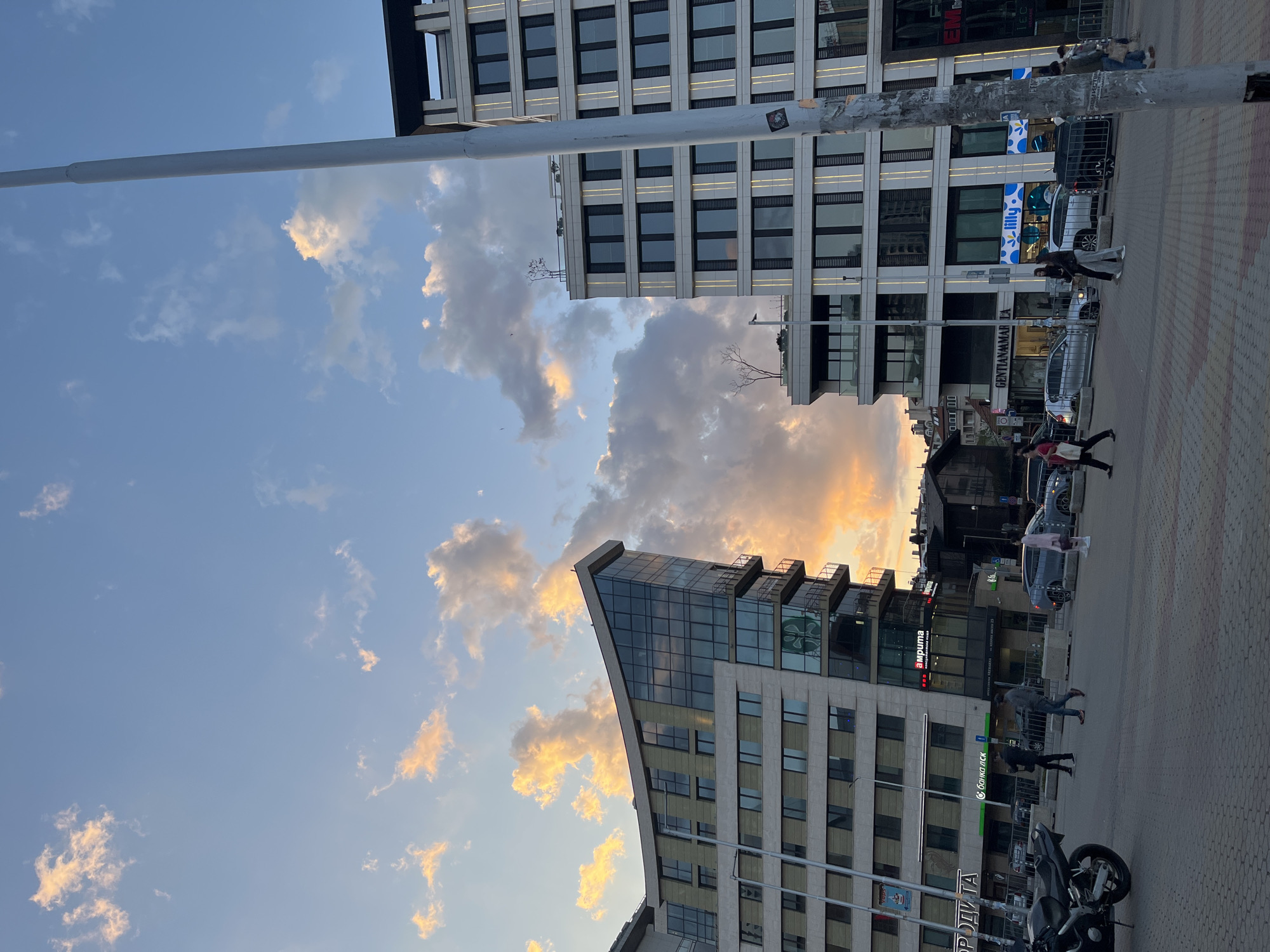 The image captures a street scene in Sofia, Bulgaria, featuring modern architecture under a sky streaked with clouds at sunset.
In the foreground, a gray pole ascends vertically along the left side. A paved walkway extends into the right side. Pedestrians walk along the street, and several vehicles are parked along the roadside.
Buildings dominate the backdrop. A modern building with glass facade and balconies rises on the right, and another more traditional, rectangular building with repetitive windows is visible in the distance. The signage on the building reads “акрима” (Akrim), “Банко ДСК” (Bank DSK), and “РОДИТА” (Rodita). The architecture, combined with the signage in Cyrillic script, place the location in Sofia, Bulgaria.
The sky is filled with clouds illuminated by a warm, golden light, suggesting either sunrise or sunset.