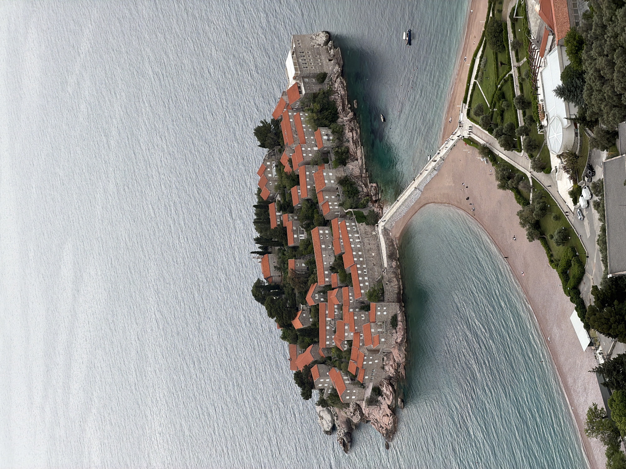 This aerial view showcases Sveti Stefan, a small islet and 5-star hotel resort on the Adriatic coast of Montenegro. The islet features a cluster of historic stone buildings with terracotta roofs, tightly packed together. A narrow isthmus connects the islet to the mainland, forming two beaches on either side. The mainland area next to the isthmus has landscaped gardens, pathways, and modern buildings. The surrounding sea appears calm, with small boats visible. The image has a somewhat rotated perspective.