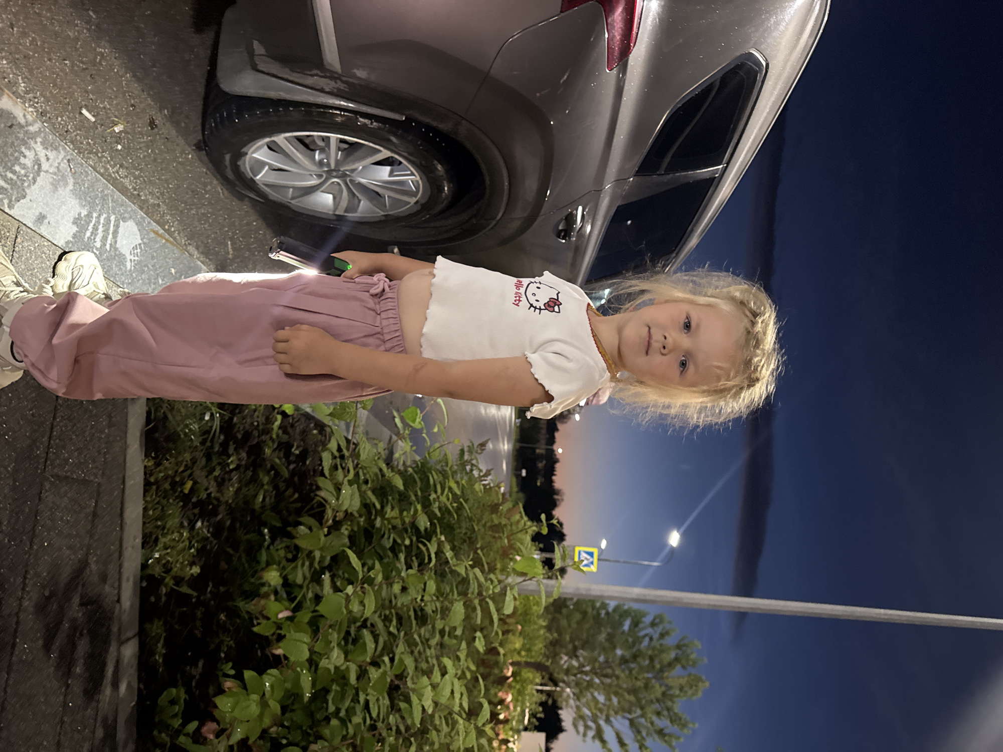 A young girl with blonde hair, wearing a white cropped shirt with a Hello Kitty graphic and pink pants, stands next to a silver SUV. She is holding a green flashlight in her right hand. The scene appears to be an outdoor parking lot at dusk, with a street and trees visible in the background. There are no identifying landmarks to suggest the city of Perm, Russia.