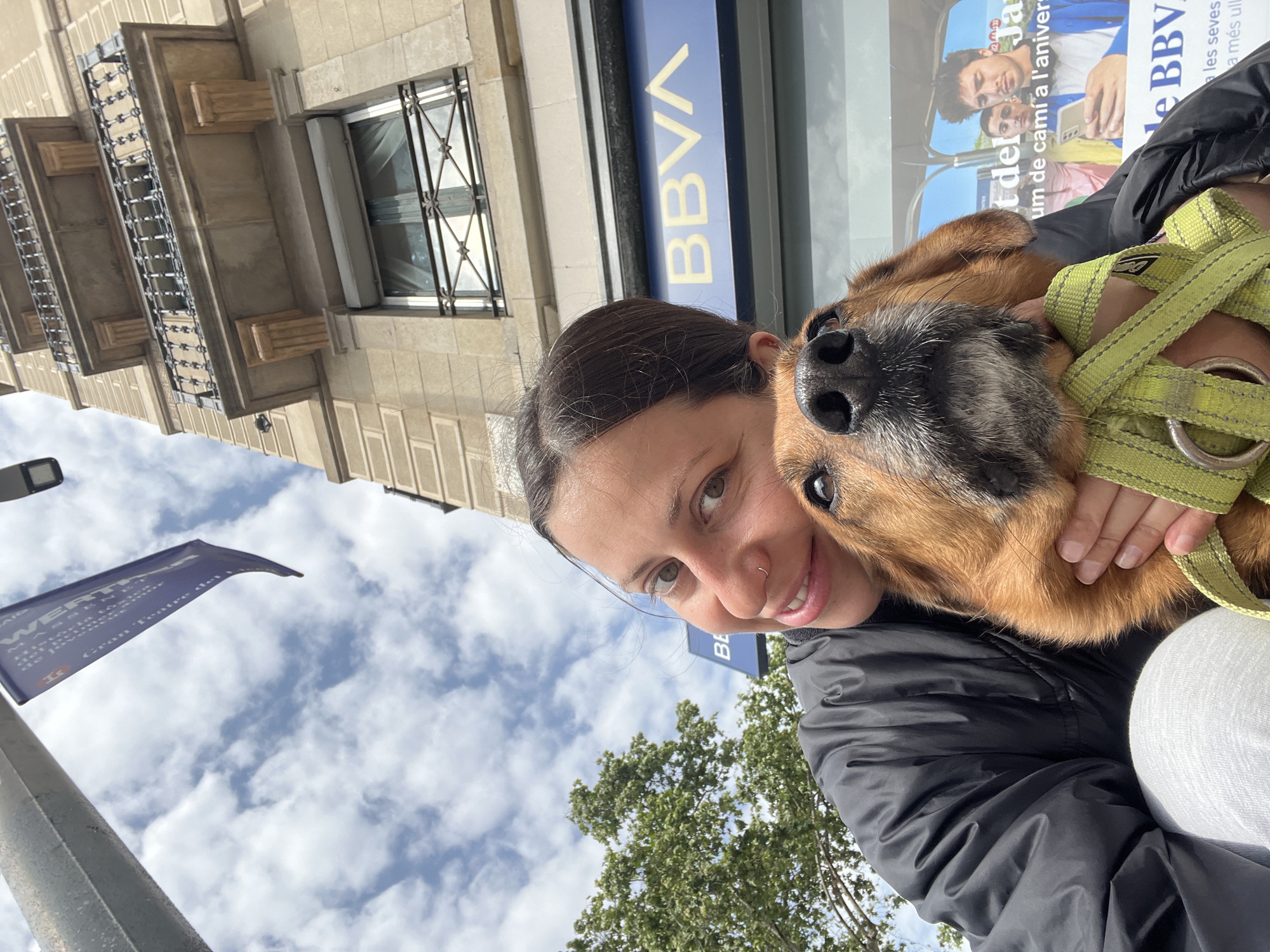 A woman holds a brown dog with a gray muzzle, appearing to take a selfie. The dog wears a green harness. Behind them is a blue BBVA bank sign and an advertisement poster with a smiling man and a text in Catalan. The background also includes a light-colored building with ornate window frames, a cloudy sky, a blue flag, and green trees. The location is likely Barcelona, Spain, based on the Catalan text on the advertisement poster.