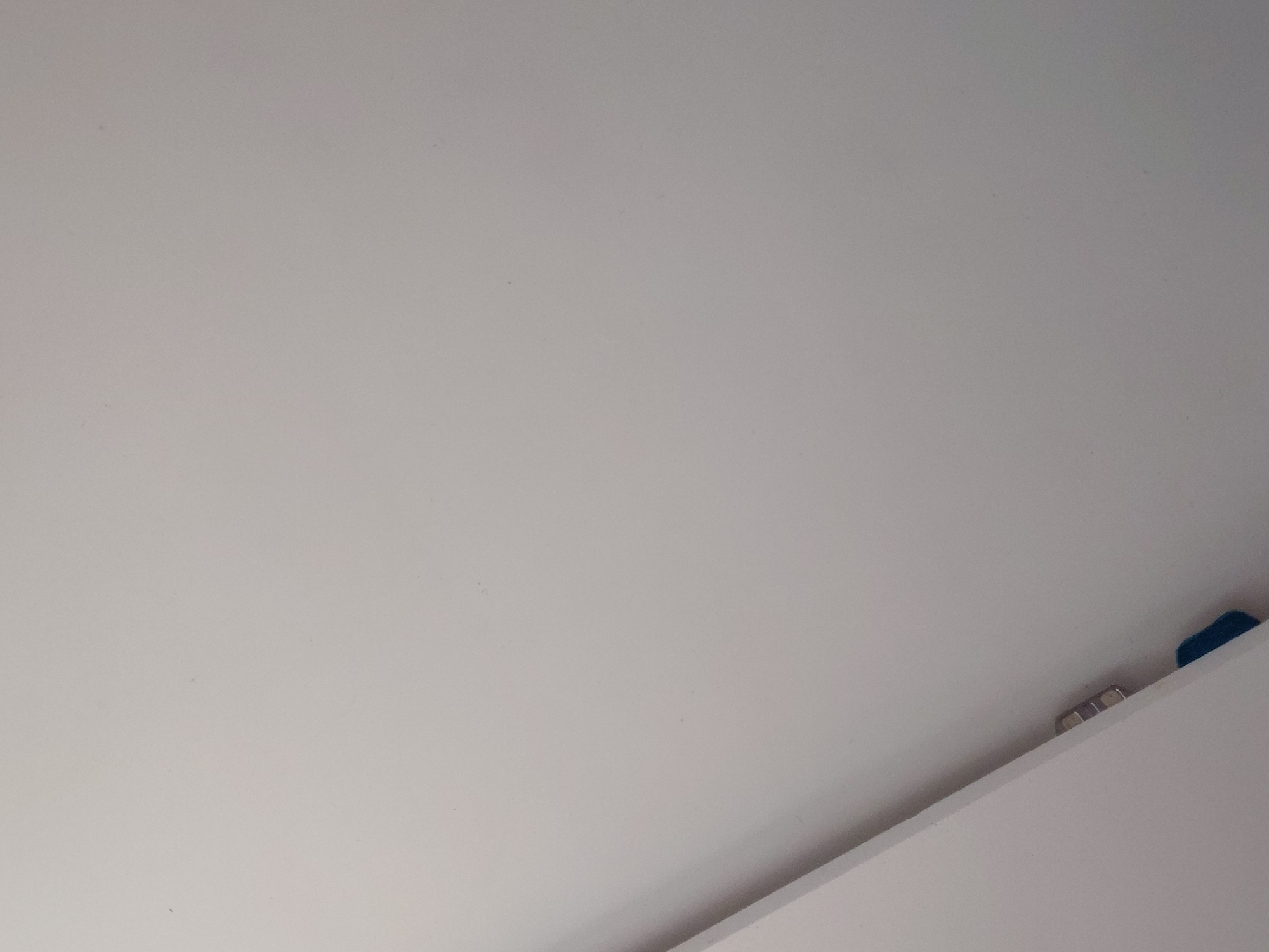 The image captures an indoor scene, primarily featuring large, unadorned surfaces. The dominant subject is a vast, smooth, light gray or off-white surface, likely a wall or ceiling, stretching diagonally across the frame from the top left to the bottom right. This surface appears uniformly painted or finished, showing minor textural imperfections and subtle variations in shading.
In the lower right portion of the image, the distinct edge of a second white surface becomes visible, creating a sharp diagonal line that suggests a corner, a joint between two panels, or the edge of a shelf or piece of furniture. Tucked into the narrow space between these two surfaces, near the bottom right corner, several small objects are present. These include two or three metallic components, possibly hinges, screws, or clips, exhibiting a silver or chrome finish. Adjacent to these, a small, vibrant blue object is partially visible, its specific function or identity indeterminate.
No people are visible in the image, and no specific activity is taking place; the scene is static, focusing on architectural or structural details. The lighting is soft and even, indicative of an interior space with diffused illumination, making the time of day and external weather conditions indiscernible. There are no location-specific cues within the image that point to Santa Ana, Venezuela. No visible text is present. The overall impression is one of clean lines and simple construction, highlighting minor functional elements within a neutral-toned setting.