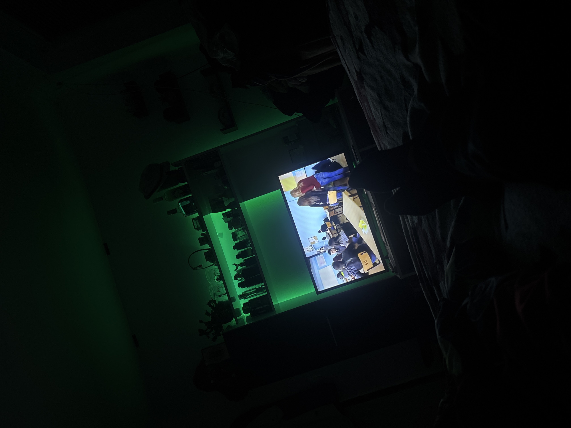 In a dimly lit room, a television screen displays a scene from what appears to be a classroom. Two women are visible on the screen, with a backdrop of desks and students. The room is illuminated by green LED strip lights placed on shelves above the television, casting an eerie glow on various objects, including bottles and figurines. The foreground is obscured by darkness, but a person's feet are visible, suggesting someone is lying in bed watching the television. The overall impression is one of a private, relaxed setting, possibly a bedroom.