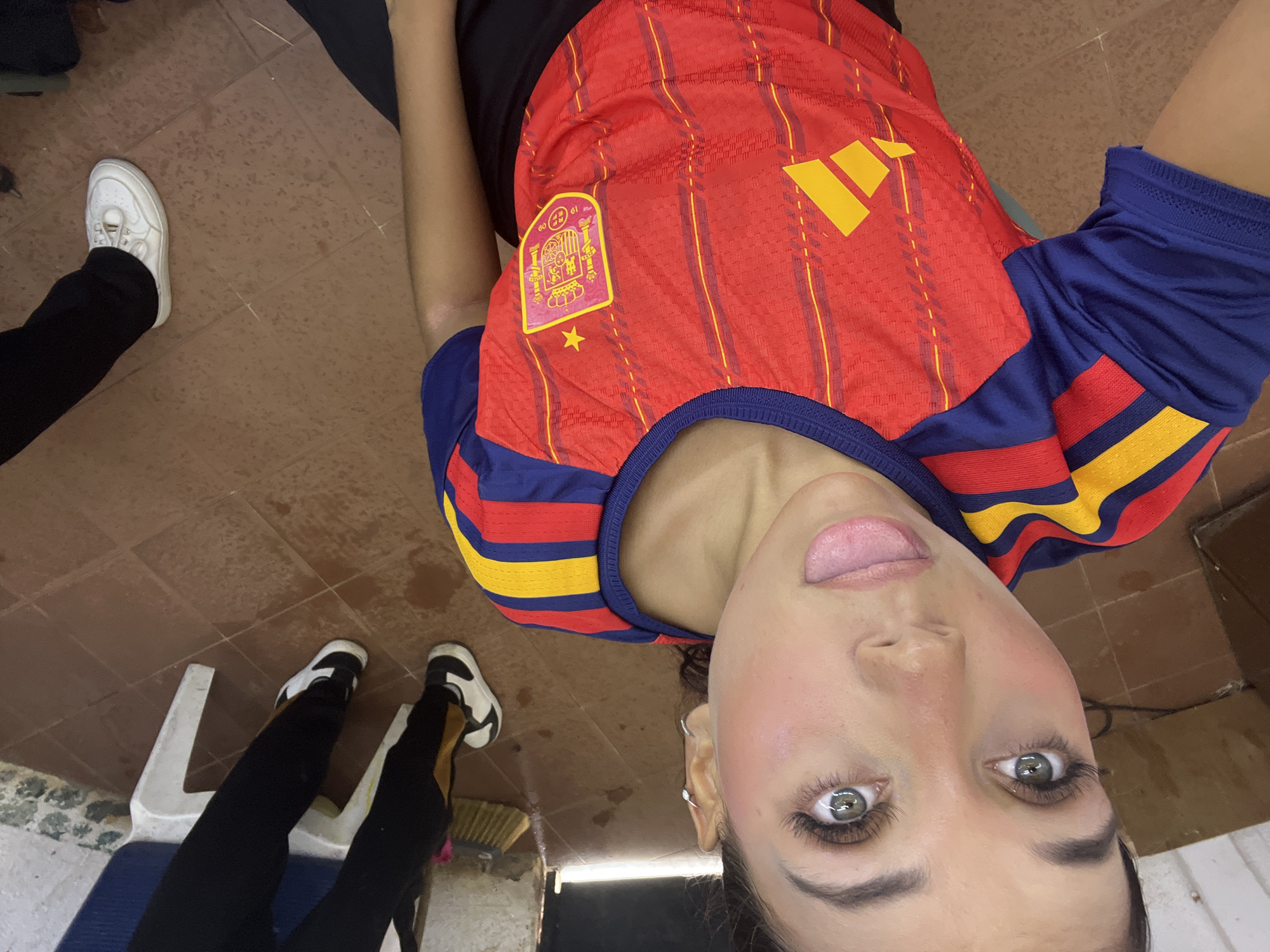 The image shows a young woman sticking her tongue out, wearing a red and blue Spanish national football team jersey. She has long eyelashes and her eyes are wide open. The background is a tiled floor, with a broom leaning against a wall. Two other people are partially visible in the background, one wearing black pants and white sneakers, and another wearing black pants. The overall scene appears to be indoors, possibly in a casual setting. There is no indication of the specific location within the city of Porlamar, Venezuela.