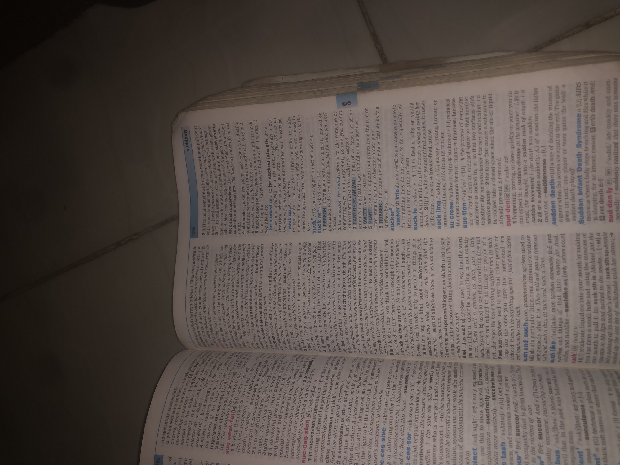 The image shows an open book lying on a surface, likely a table. The book is a dictionary or reference book, as indicated by the entries for words such as "suddenly," "suck," "sucker," and "succulent." The pages are filled with text and some small illustrations. The lighting suggests it may be nighttime or a dimly lit indoor space, casting shadows on the pages. The book is the primary subject of the image, and there is no visible connection to Jalingo, Nigeria, or any specific city. There are no people, actions, or interactions depicted.