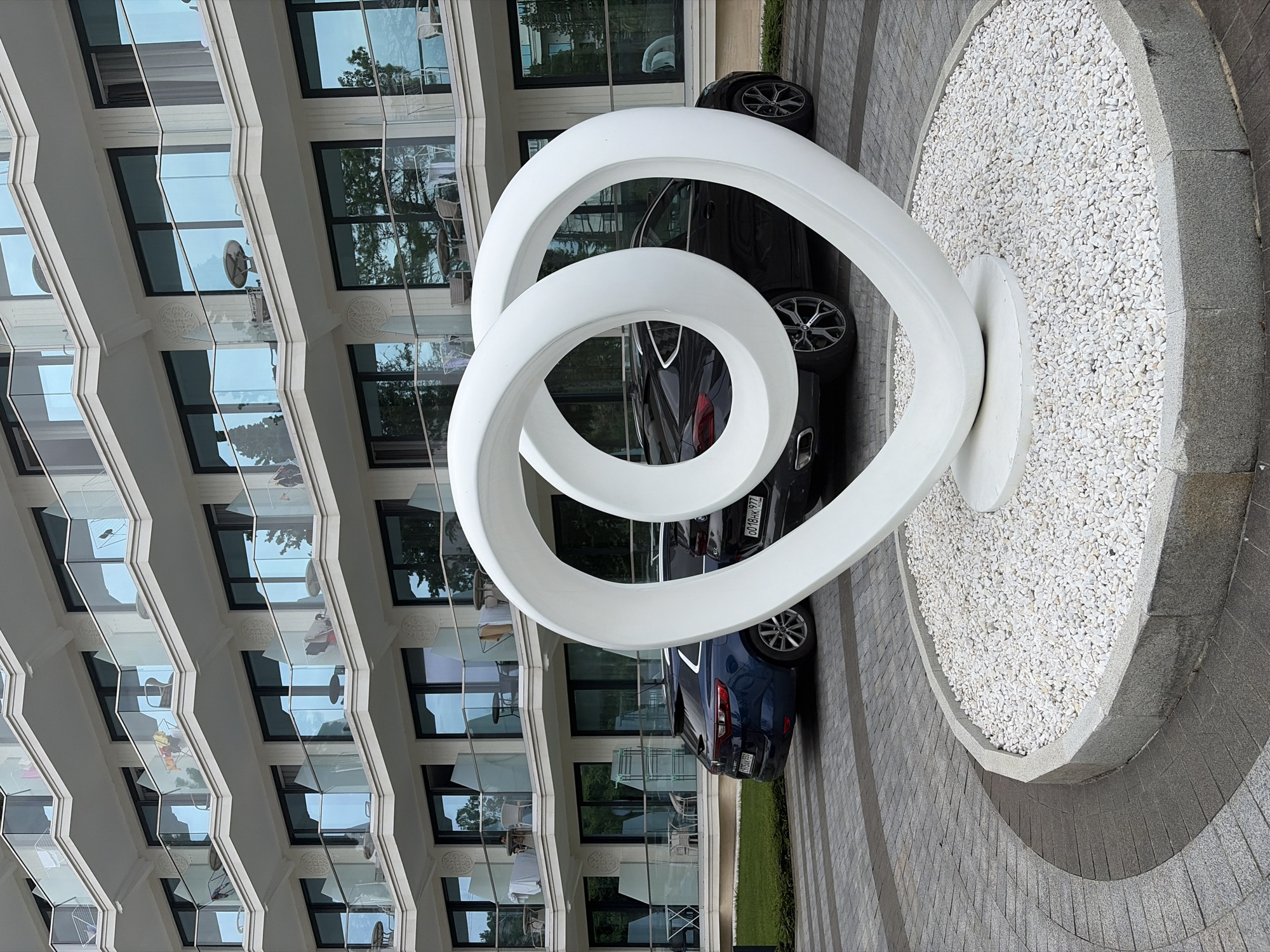 The image shows a modern apartment building with multiple balconies overlooking a courtyard. In the foreground, a large white abstract sculpture shaped like an infinity symbol or intertwined loops is positioned in a circular garden bed filled with white gravel. Two cars, a dark blue SUV and a black SUV, are parked in the courtyard. The building's facade is white with cream-colored trim and glass balconies. The ground is paved with interlocking gray stones and cobblestones. The scene appears to be in Sochi, Russia, given the architectural style and the presence of a modern residential complex. There are no people visible in the image.