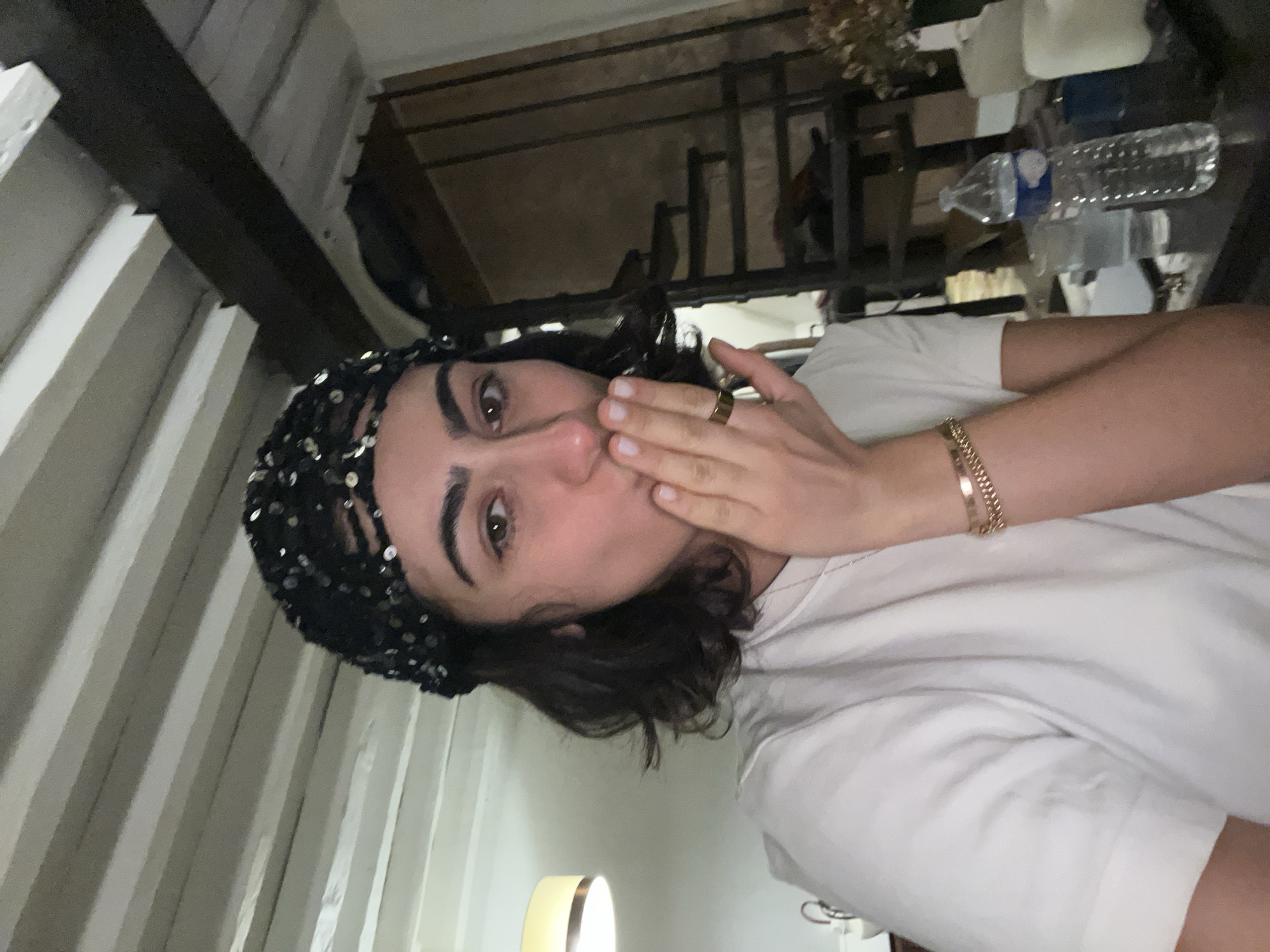 A person with dark hair, wearing a black sequined head covering, is positioned at an angle, appearing to blow a kiss. They are wearing a gold ring and a gold bracelet. The person is wearing a white t-shirt. In the background, there is a glimpse of a room with a metal shelf, water bottles, and some dried flowers. The setting resembles a Parisian apartment with corrugated metal walls and a wooden ceiling beam visible on the left. The lighting is dim and artificial, with a lamp visible in the lower center of the frame.