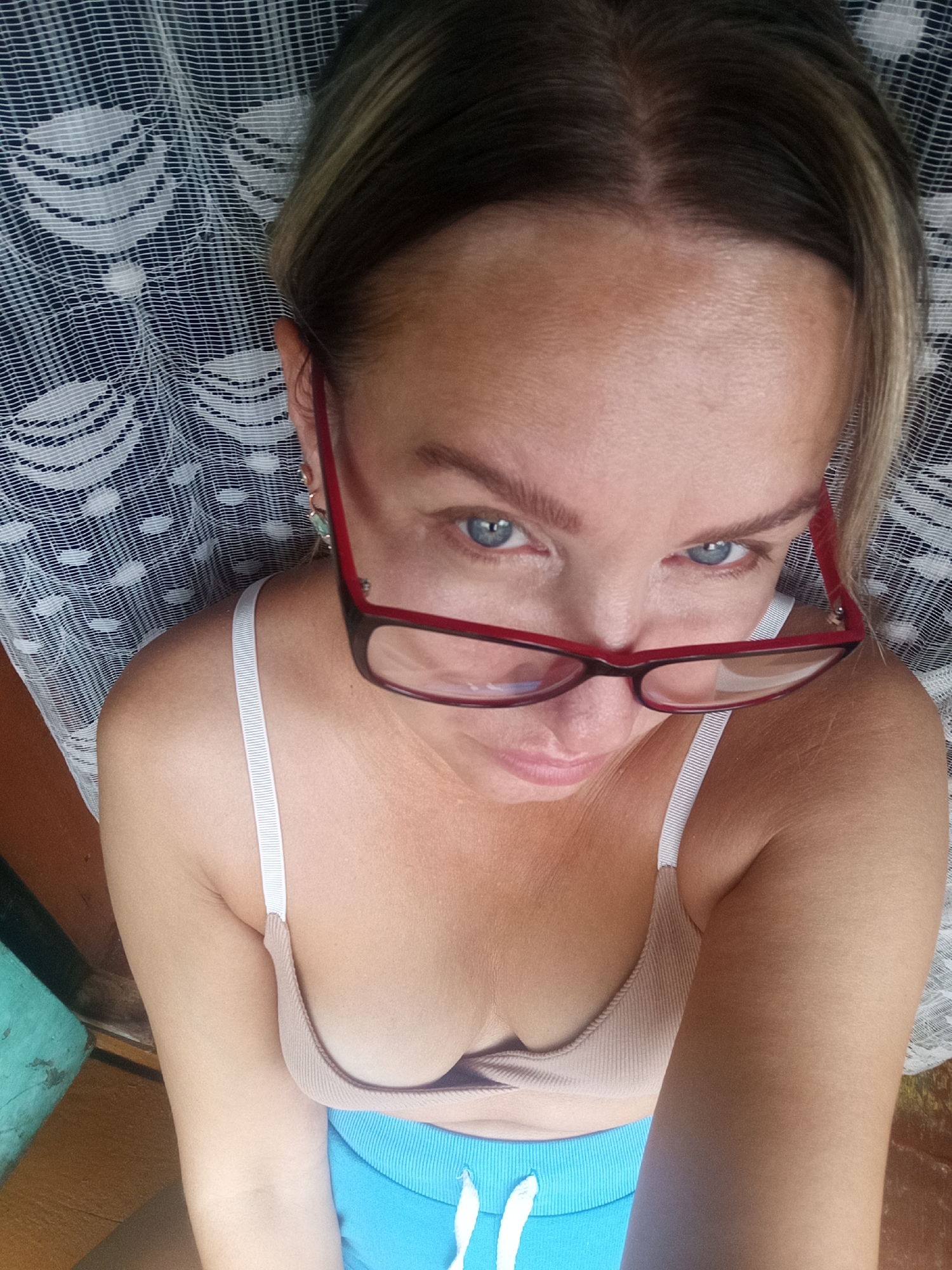 A woman wearing red-rimmed glasses, a tan bralette, and blue shorts poses for a selfie. She has blonde hair and blue eyes. The background consists of a white lace curtain and a wooden surface. The setting appears to be indoors, possibly in a home.