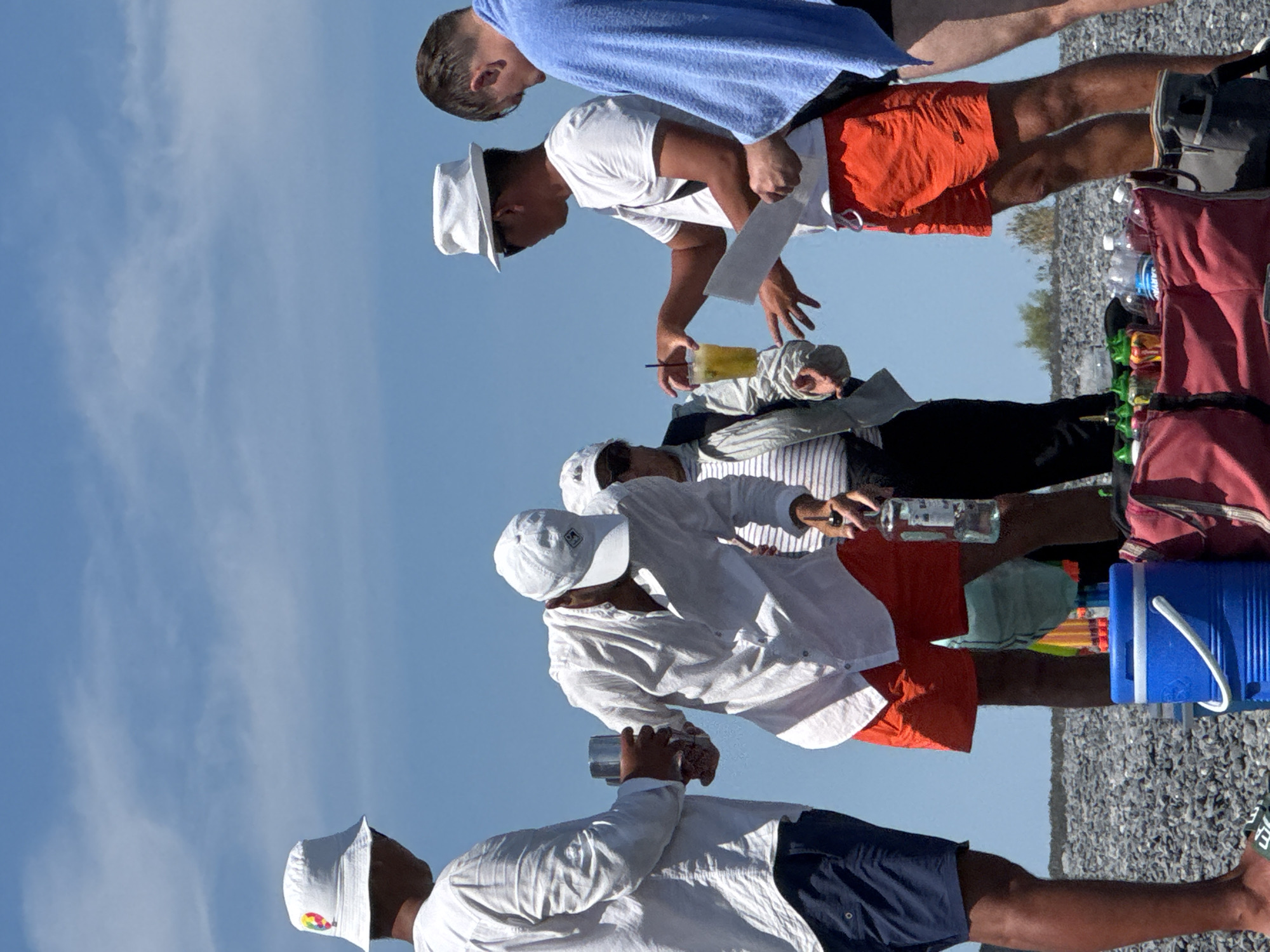 The image depicts a group of five individuals standing on a rocky surface under a clear blue sky. The group appears to be engaged in a casual outdoor gathering, possibly a picnic or a break during an activity.
In the foreground, a person on the far left, wearing a white bucket hat and a white long-sleeved shirt, is holding a metallic cup. To their right, another individual in a white short-sleeved shirt and orange shorts is seen bending down, holding a bottle in their left hand and a metallic cup in their right. Next to them, a person in a light-colored jacket and striped shirt is partially visible, also holding a bottle.
Further to the right, a person wearing a white hat and sunglasses is holding a clear plastic cup with a straw containing a yellow beverage, and a paper in their left hand. To their right, another individual is draped in a blue towel over their shoulder, and is wearing black shorts.
In the background, a blue cooler with a white handle is positioned on the ground, alongside a red bag and a gray bag. Several colorful items, possibly party favors or decorations, are visible near the bags. The ground is covered with small, gray stones, and some sparse vegetation can be seen in the distance. The overall setting suggests an outdoor recreational area, likely a beach or a riverside, in the vicinity of Ақши, Kazakhstan.