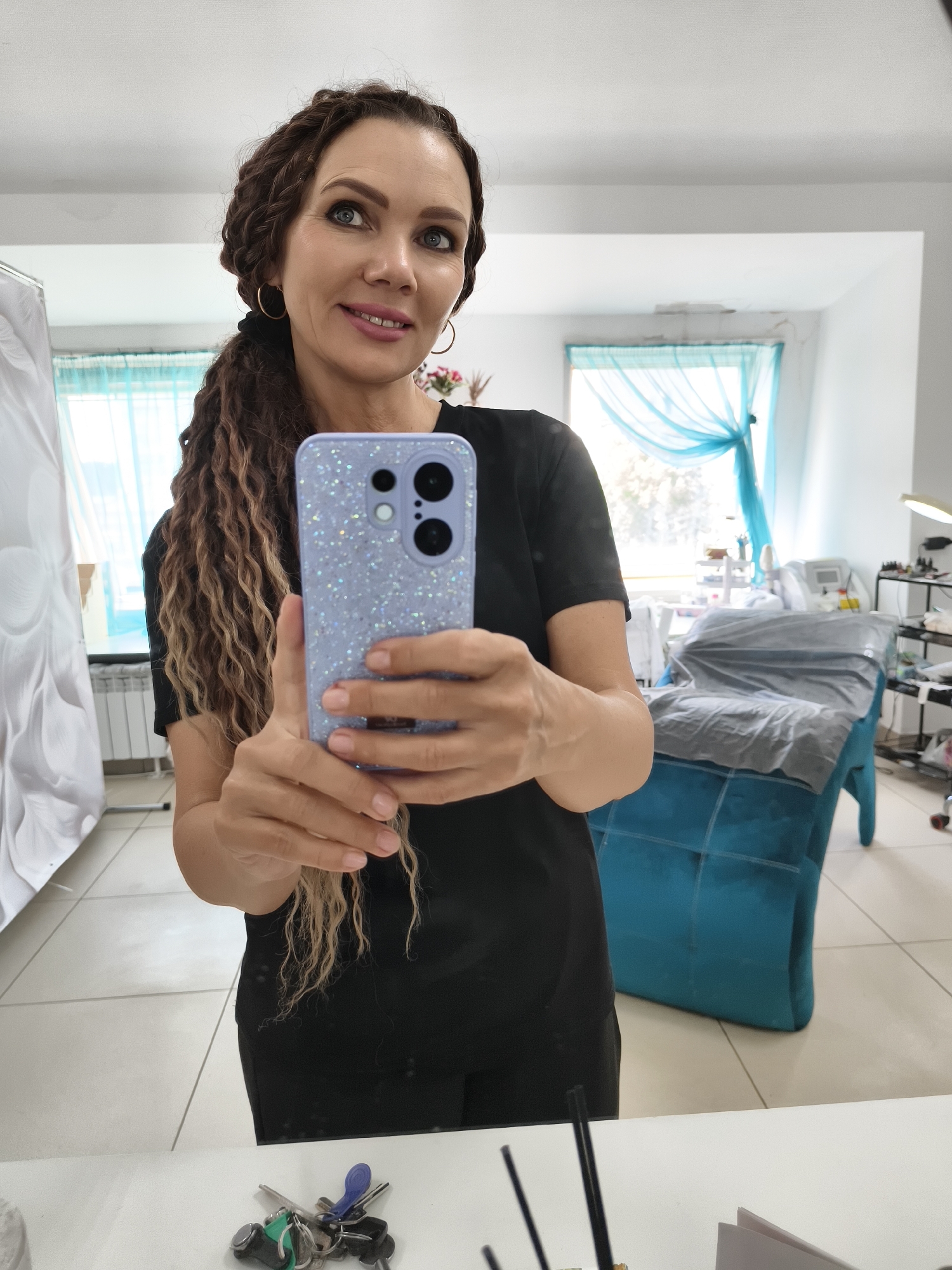 In a well-lit room, a woman with long, braided brown hair tied in a ponytail takes a mirror selfie. She is wearing a black t-shirt and holding a light purple phone with a glittery case. The phone's camera is pointed towards the mirror, capturing her reflection. Her expression is a slight smile as she looks at the phone.
In the background, the room appears to be a salon or spa. A large, turquoise tufted couch or treatment bed is visible, covered with a grey sheet. Behind the bed, a window with sheer blue curtains allows natural light to enter. Various salon equipment, including shelves with bottles and a lamp, are organized in the space. The floor is tiled with white and grey tiles. On a white surface in the foreground, a set of keys is scattered, along with a few other small objects. The overall impression is of a professional space ready for client services.