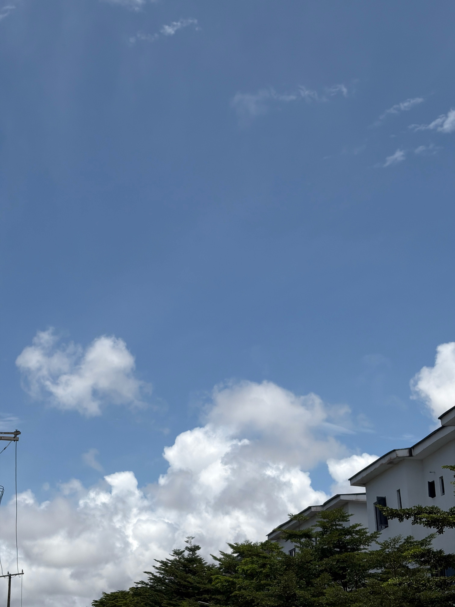 The image depicts a clear blue sky with scattered white cumulus clouds over a line of white modern houses with peaked roofs, partially obscured by lush green trees in the foreground. A utility pole with wires is visible on the left side of the frame. The scene appears to be an outdoor residential area in Igboefon, Nigeria. There are no people visible in the image, and no actions or interactions are taking place. The environment is a daytime outdoor setting characterized by fair weather conditions.