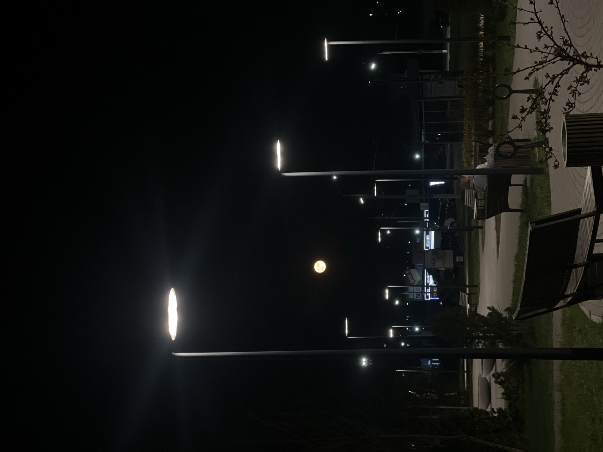 The image depicts a night scene in what appears to be a park or public space in the city of Chaikovsky, Russia. Several modern-style streetlights illuminate a pathway lined with benches and some sparse vegetation. A full moon is visible in the sky between the streetlights. One person can be seen sitting on a bench in the distance. Further down the pathway are businesses, and possibly traffic.