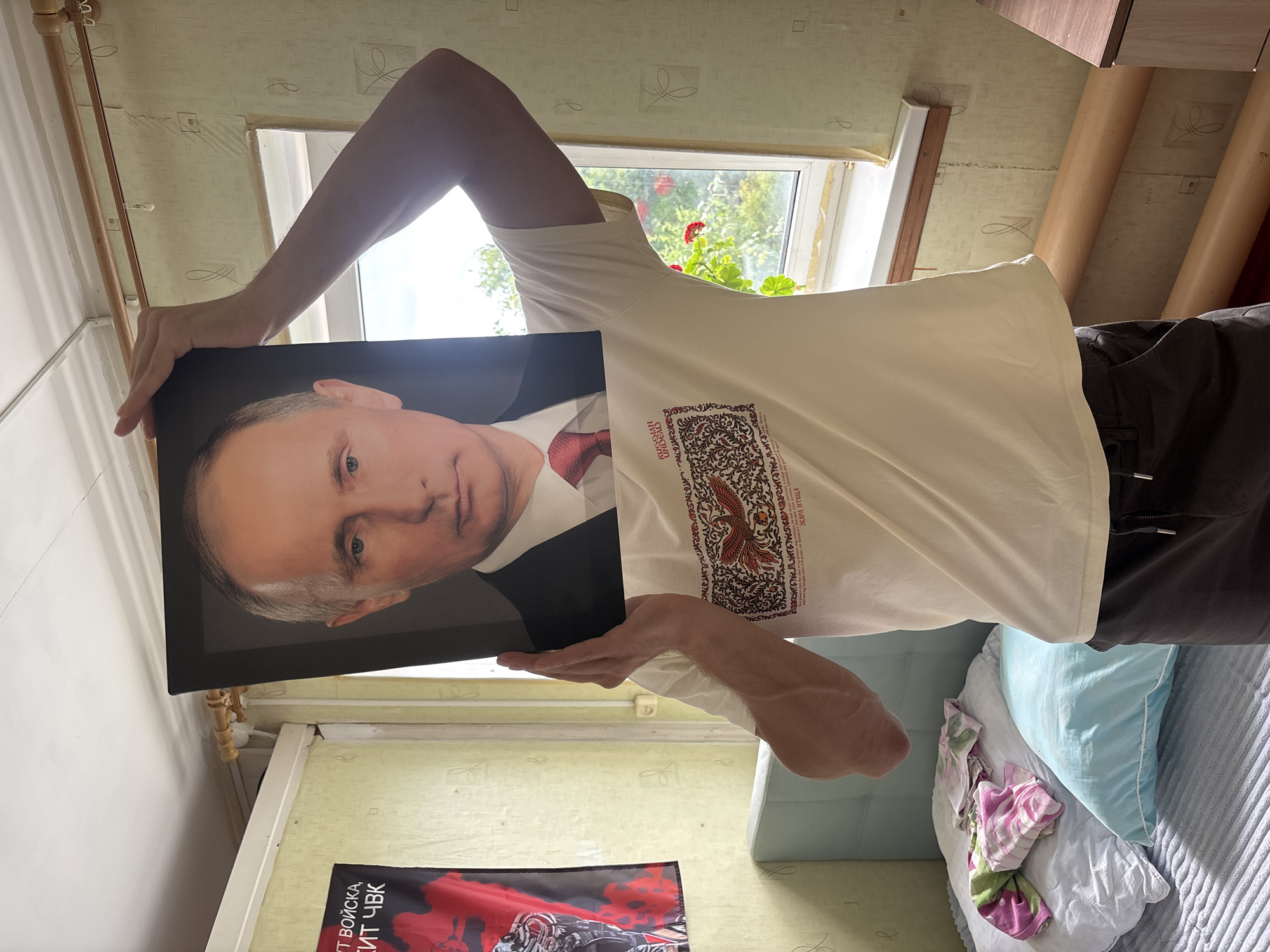 A person holds a framed portrait of Vladimir Putin in front of their face. The portrait is of Putin's head and shoulders, showing him wearing a dark suit and a red tie. The person is wearing a light-colored t-shirt with a graphic design on the front, and dark pants. They are standing in a room with a bed on the left side, partially covered with bedding and pillows. A red and black flag with Cyrillic text is hanging on the wall to the left of the person. A window with a sill and curtains is visible on the right. The room appears to be a bedroom, with walls covered in patterned wallpaper and a radiator. The overall scene is casual and indoors. The image does not provide enough information to determine the specific location within Krasnogorskoye, Russia.