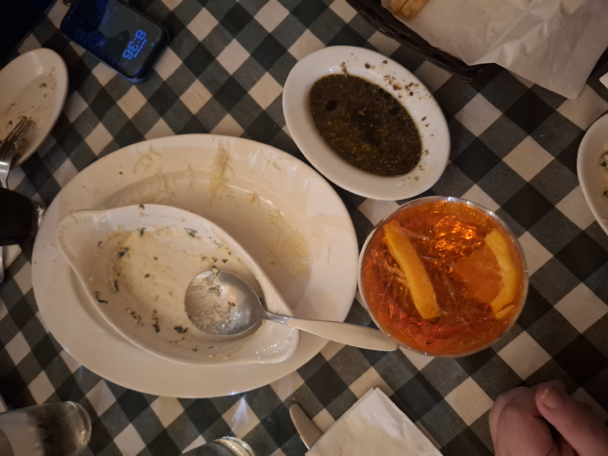 A close-up overhead shot captures a partially cleared restaurant table, covered with a green and white checkered tablecloth. The scene features several plates and serving dishes, most of which appear to have been used. One larger plate has remnants of food and is partially obscured by a smaller dish with a spoon resting inside. Another small, oval-shaped dish contains a dark green sauce or dip. A glass filled with a vibrant orange liquid, possibly a cocktail or juice, sits to the right of the large plate and appears largely untouched. A phone is visible in the upper left corner, displaying a time of 8:36. Portions of a bread basket, napkins, cutlery, and a partially visible hand are also present on the table. The overall impression is that a meal has been partially consumed, and the setting is likely a restaurant environment. While the image provides context, pinpointing a precise location within Naucalpan de Juárez, Mexico, is impossible without additional clues.