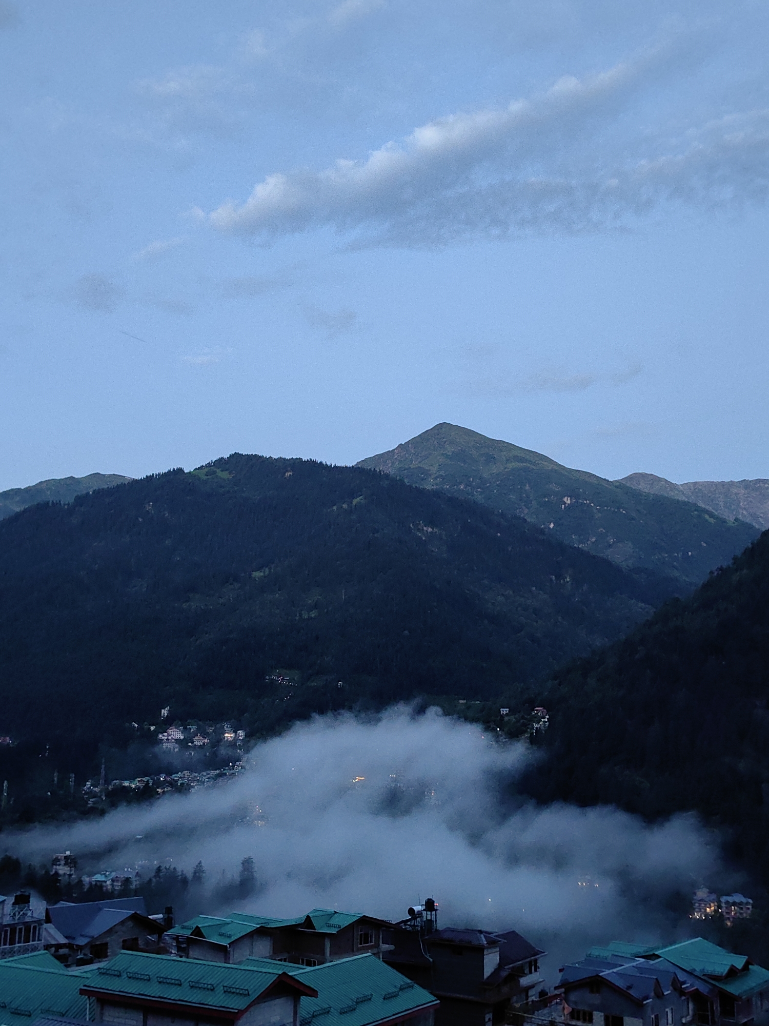 In a twilight mountain landscape, a village is nestled amidst dense, dark green coniferous forests that blanket the steep slopes. The sky above is a soft, hazy blue with scattered, wispy clouds, suggesting either early morning or late evening. A thick layer of white mist and fog rolls through the valley, partially obscuring the buildings of the village, creating an ethereal and atmospheric effect. The architecture of the village is visible in clusters, with buildings featuring dark roofs, some with a slight bluish-green hue. The foreground shows the rooftops of several houses, some with visible solar water heaters. The scene is serene, with no visible people or animals, emphasizing the natural beauty of the mountainous terrain and the quiet existence of the settlement. This is likely the village of Vashisht, India, known for its hot springs and scenic location in the Himalayas.