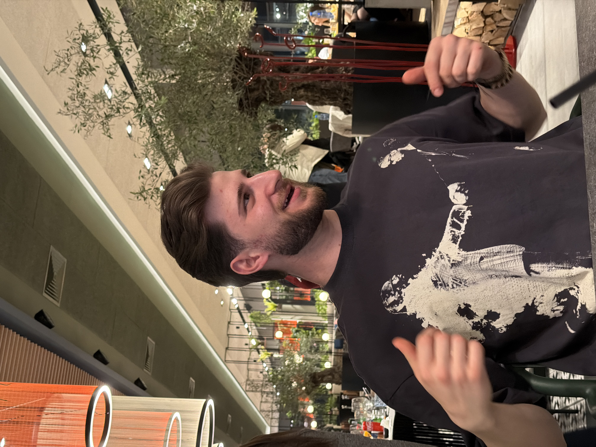 The scene depicts a young adult male seated in a modern restaurant or bar establishment, located in Plovdiv, Bulgaria. The male, with light skin, brown hair styled upwards, and a trimmed brown beard and mustache, is smiling broadly, teeth visible, and looking towards the right side of the frame. He is wearing a dark charcoal grey short-sleeved t-shirt featuring a large white graphic print of an abstract, splattered human-like figure. Both of his hands are raised slightly, with thumbs extended upwards in a positive gesture.
The environment is characterized by contemporary decor. The ceiling incorporates sections of light-colored wooden slats alongside flat dark grey panels equipped with rectangular ventilation grilles and recessed spot lighting. Prominent orange and light grey cylindrical pendant lamps, each featuring internal ring lighting, hang in the upper left of the frame. Several large, potted olive trees with gnarled trunks and green foliage are distributed throughout the background, serving as significant decorative elements. Slender red decorative rods are arranged vertically near one of the olive trees. A decorative stack of light-colored firewood logs is visible on the right side behind the subject.
In the mid-ground and background, other patrons are partially visible; a person in a light-colored jacket is obscured by an olive tree, and another individual is seated at a distant table on the far right. A table to the left in the background is set with glassware and bottles, including a red Coca-Cola bottle. The foreground shows a dark, smooth table surface, with a dark straw or stirring stick visible. The overall lighting is warm, contributing to a comfortable evening ambiance.