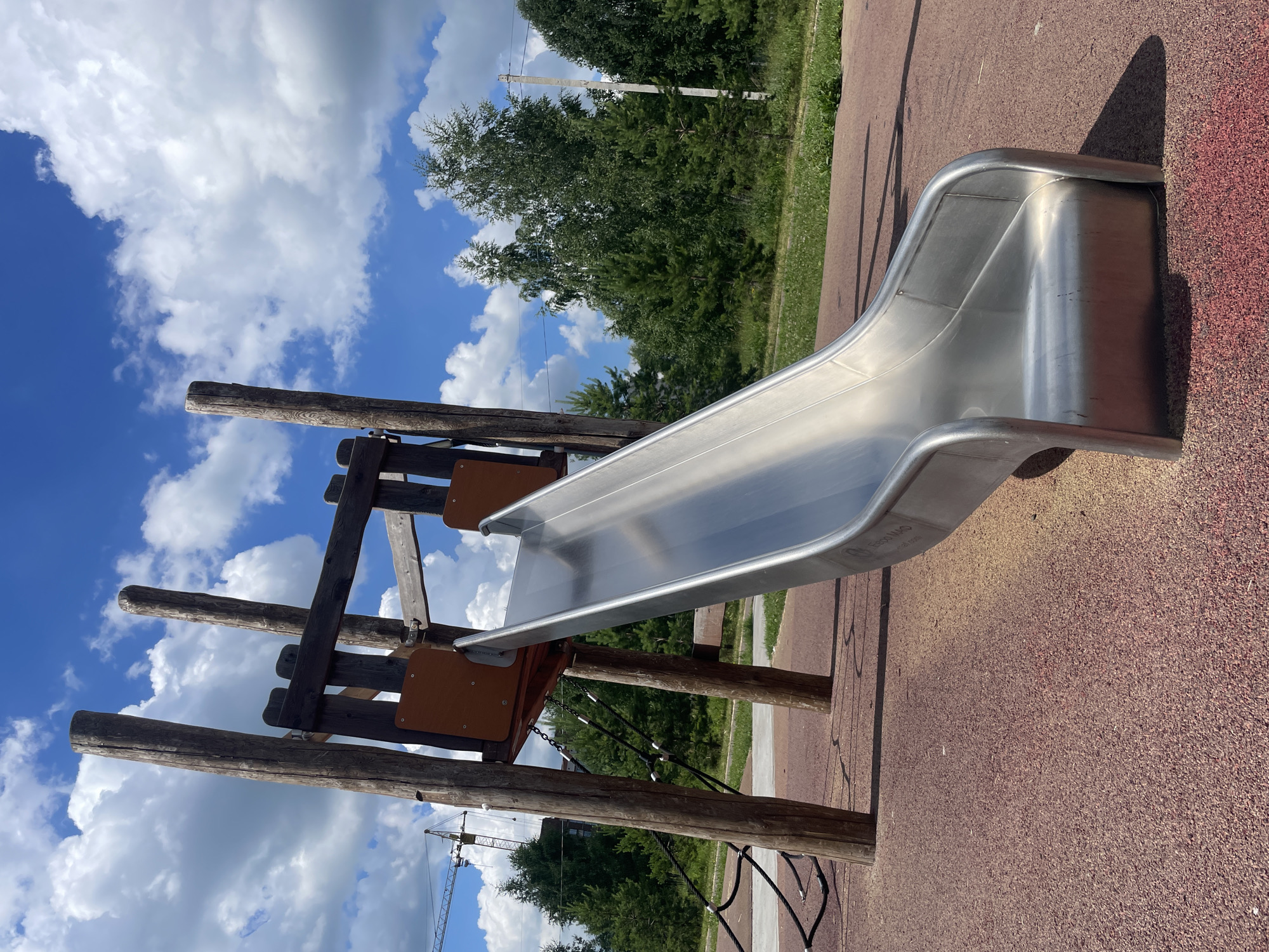 This media file captures a modern metal slide with a wooden frame on a playground. The slide is silver and curves downwards, with rounded edges. The supporting structure is made of thick, rough-hewn logs, connected by dark brown wooden planks and metal fixtures. There are also some brown wooden panels attached to the structure, possibly for climbing or resting. Black ropes, secured by metal clasps, form a net-like barrier or climbing element below the slide's entrance.
The playground surface is a reddish-brown, textured material, likely a safety surfacing common in children's play areas. In the background, lush green trees and foliage are visible, indicating an outdoor setting. The sky is a vibrant blue, dotted with fluffy white clouds, suggesting a pleasant, possibly warm day. A tall white pole with wires, likely a utility pole, is also visible among the trees. In the far distance, a construction crane can be seen, hinting at ongoing development in the area.
The image is taken from a low angle, emphasizing the slide and its structure. The lighting indicates it's daytime, with shadows cast by the slide and the wooden supports, suggesting the sun is at an angle. There is no visible text in the image, except for a small inscription on the slide itself, which is not legible enough to be read. The overall impression is one of a well-maintained outdoor playground in a natural or semi-urban environment on a clear day.
