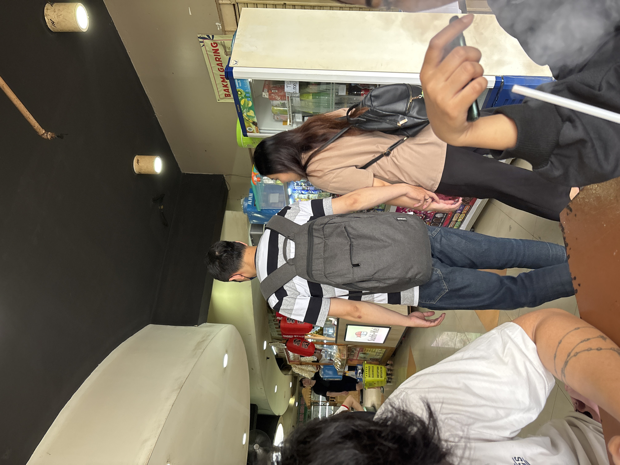 A couple is walking hand-in-hand through a market in North Jakarta, Indonesia. The man, wearing a striped t-shirt and backpack, walks in front of the woman, who is wearing a brown t-shirt and a black backpack. In the background, a man in a black t-shirt is standing behind a food stall. The market appears to be outdoors, with a dark ceiling and fluorescent lighting. There are various food stalls and refrigerators visible, selling drinks and snacks. The overall atmosphere is casual and busy, suggesting a typical day at a local market.