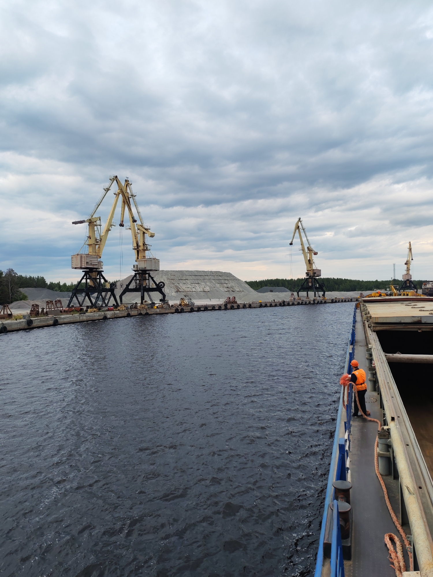 The image depicts a port scene in Shal'skii, Russia, under a cloudy sky. In the foreground, the edge of a large cargo ship is visible, with a person in an orange safety vest holding a thick rope. The ship's deck is dark and weathered.
Beyond the ship, a dark body of water stretches towards a concrete pier. Along the pier, several large yellow cranes are positioned. Two prominent cranes dominate the left side of the frame, standing tall with their booms extended. Behind them, a vast pile of gray aggregate material is stacked. Further down the pier, two more cranes are visible, smaller in scale.
The background features a dense line of green trees, suggesting a forested area bordering the port. The overall atmosphere is industrial and functional, with a focus on the logistics of cargo handling. The water is choppy, indicating a potentially windy or active marine environment.