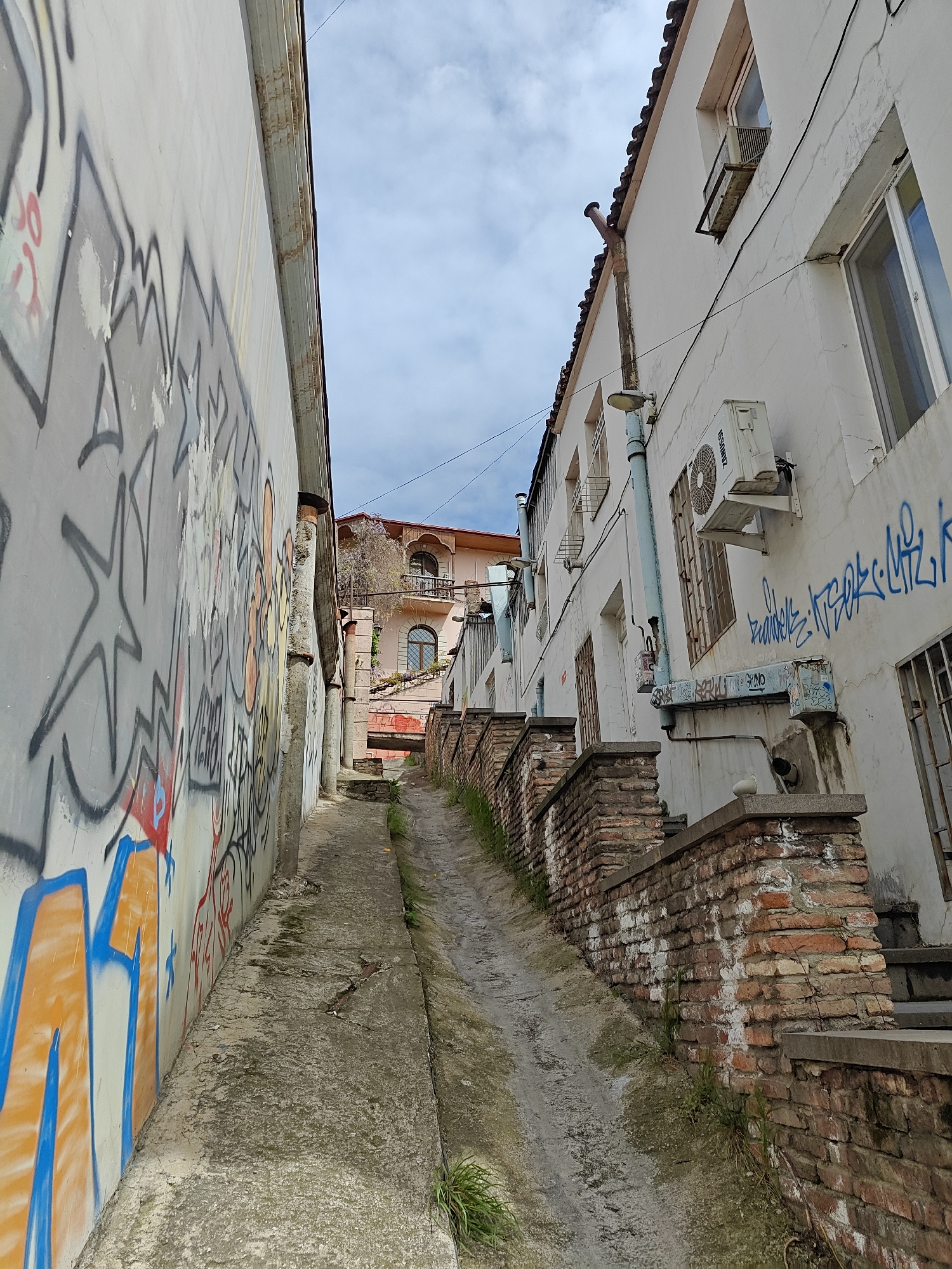 The image depicts a narrow alleyway in the city of Tbilisi, Georgia. The alley is paved with worn concrete, and a narrow drainage channel runs along its center. The alley ascends uphill. The wall on the left is covered in extensive graffiti of various colors, while the wall on the right is the side of a white building with windows, air conditioning units, and graffiti. A low brick wall runs along the base of the building. In the distance, another building with a reddish facade is visible, framed by the alleyway. The sky above is partly cloudy.
