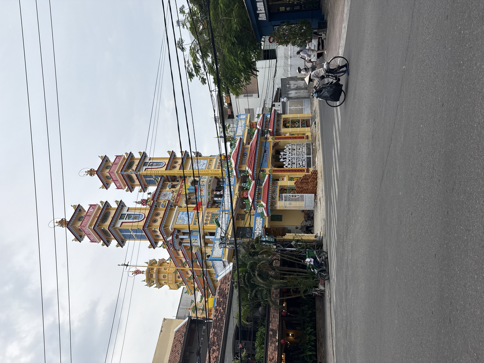 The image captures a street scene in Nha Trang Ward, Vietnam. A tall, multi-tiered, brightly colored building with yellow, blue, and red accents dominates the left side of the frame. Overhead power lines crisscross the sky above the building. On the right, a street runs alongside the building, with two people visible. One person, wearing a conical hat, is cycling down the road. Another person is walking along the sidewalk. A parked scooter and other parked bicycles are visible near the sidewalk. Other buildings and some greenery are in the background.