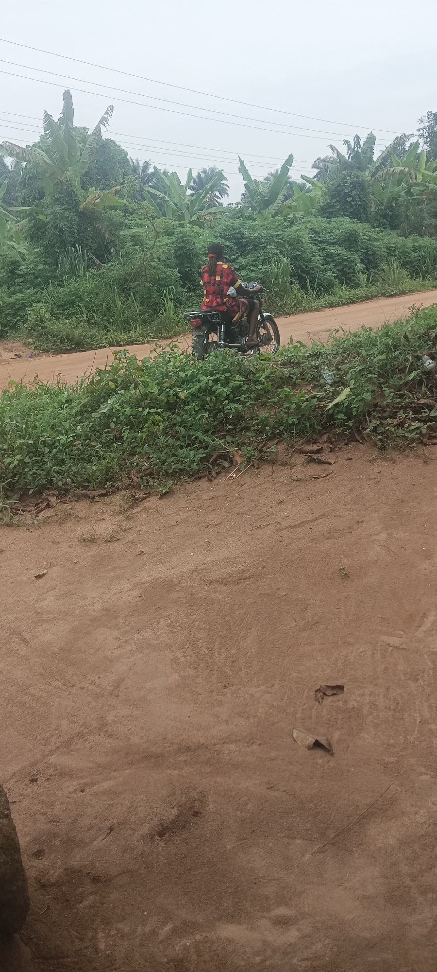 A person in a red plaid shirt rides a motorcycle on a dirt road in Amauzu, Nigeria. The environment is lush, with abundant green vegetation and banana trees visible in the background. Power lines are strung overhead. The foreground shows the dirt road surface with scattered leaves.