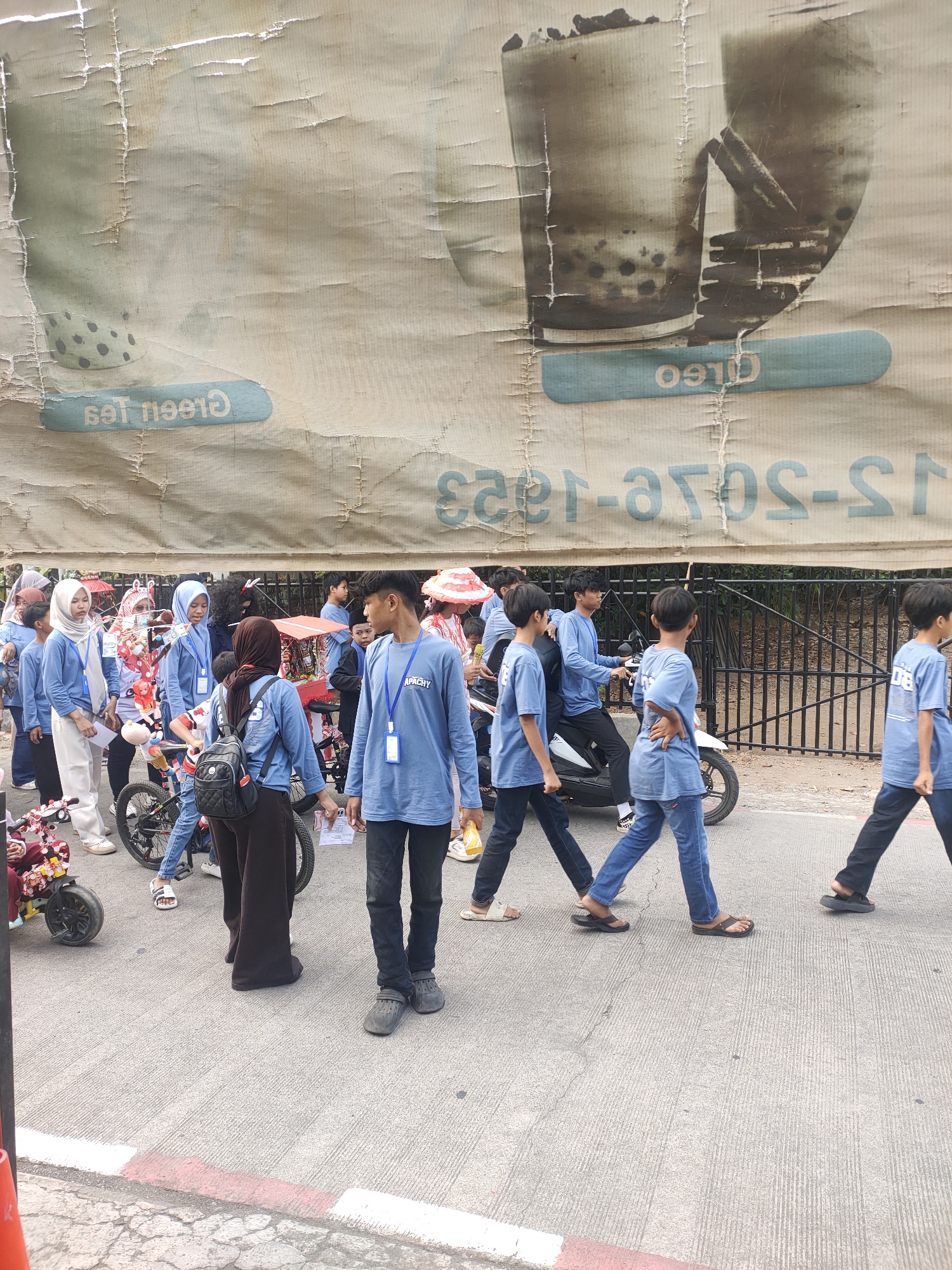 In Subang, Indonesia, a group of youths, predominantly wearing blue shirts, are participating in a street event. The youths are walking on a paved road, with a large, weathered banner hanging overhead. The banner displays faded graphics of a beverage, possibly bubble tea, and text in both English and Indonesian, reading "Green Tea" and "Oreo" horizontally and "23-JOS-S-I JOS" vertically. The participants are diverse in age, with some appearing to be teenagers and others younger. Some are walking, while others are near bicycles and a scooter, suggesting a procession or parade. Several individuals are wearing lanyards with identification badges. The environment appears to be outdoors, possibly a public street or square, with trees and a fence visible in the background. The overall scene depicts a lively community event.