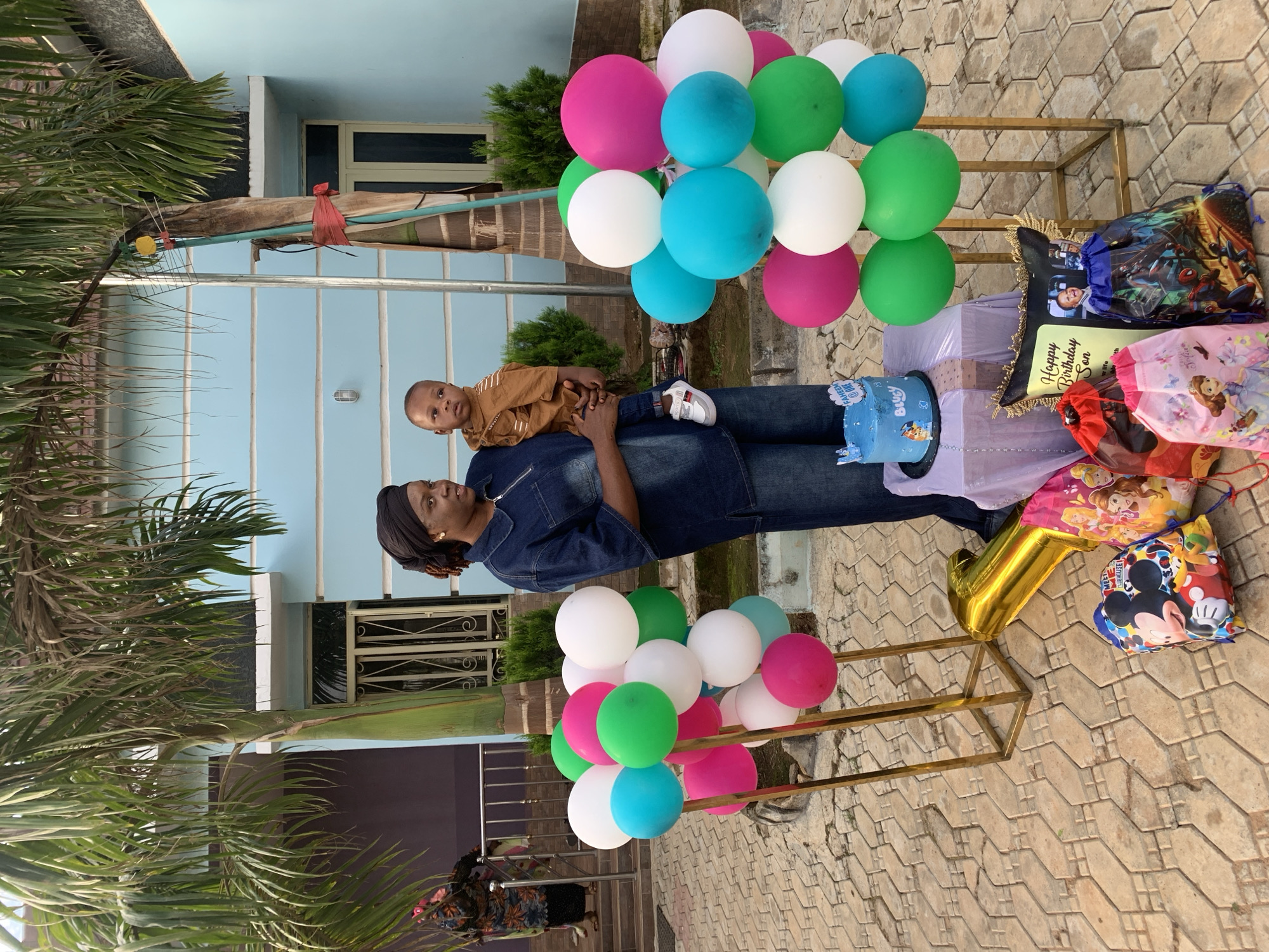 This image captures an outdoor scene, likely a celebratory event, in the city of Jos, Nigeria. A dark-skinned woman, wearing a dark blue denim top and trousers with a black headwrap, stands slightly to the right of the frame, holding an infant. The infant, wearing a brown shirt and blue jeans, faces forward but looks to its right. To the left, a partially visible woman in a patterned dress ascends a staircase towards a building.
The foreground features various birthday decorations. Two tall, golden metal stands display clusters of balloons in hot pink, bright green, white, and light blue. Between these stands, a small table covered with light purple and white fabric holds a round, light blue cake inscribed with "FAHEEM @ ONE" and featuring a "Bluey" character. A large, golden foil balloon shaped like the number "1" rests on the light-colored paved ground in front of the table. Several drawstring bags with cartoon characters (Mickey Mouse, Disney Princesses, Spider-Man, Sofia the First) are arranged on the pavement, alongside a framed "Happy Birthday Son" item featuring a baby's photograph.
In the background, a light blue building with white trim and barred windows is visible, partially obscured by two large palm trees. The ground is paved with tan-colored interlocking tiles.