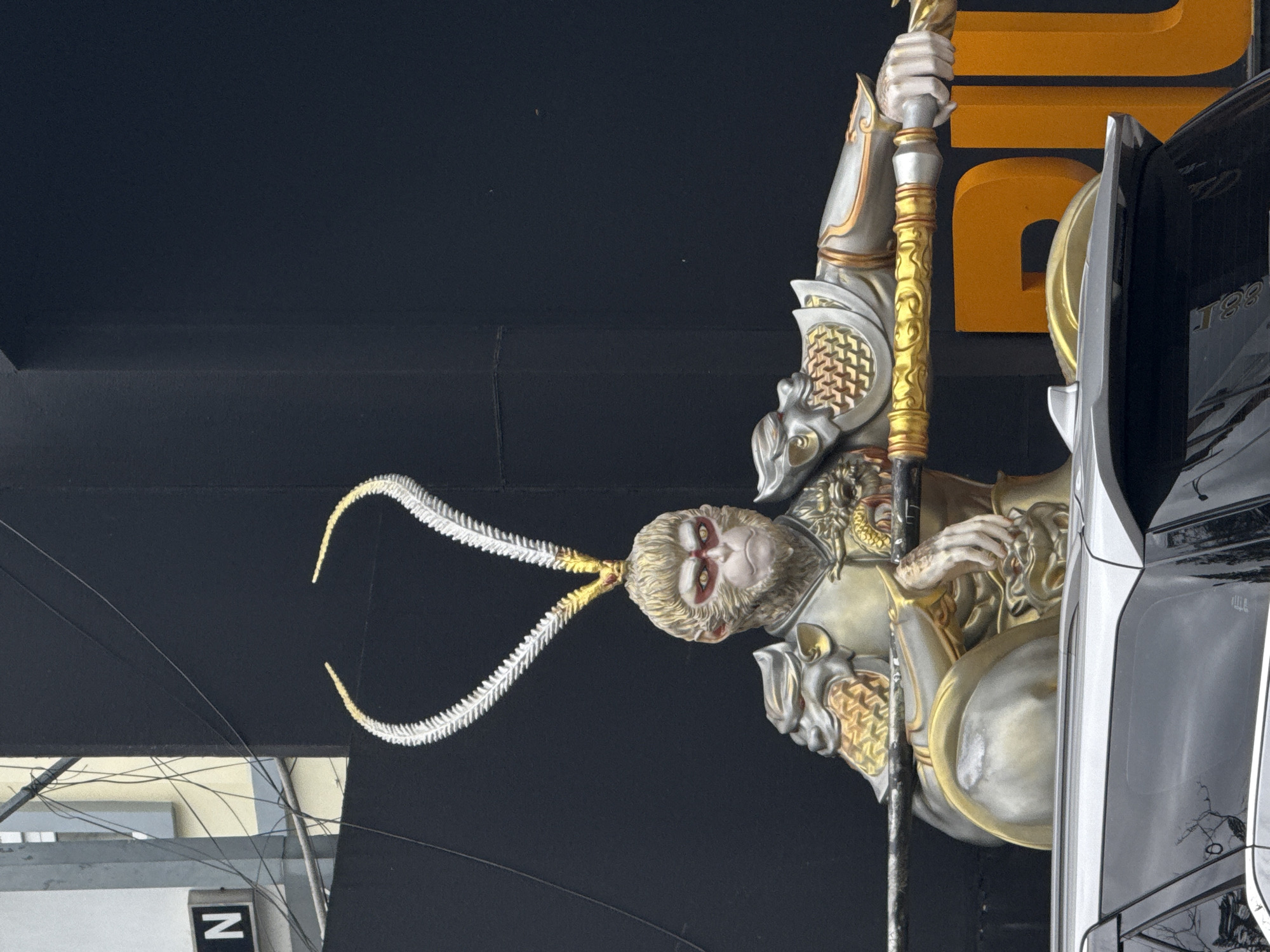 An outdoor, eye-level shot captures a large statue of the Monkey King from the Chinese epic "Journey to the West." The statue is depicted as a muscular, armored figure with golden and silver armor, resting on what appears to be a long staff. The Monkey King has distinct facial features with red around its eyes and two large, feathery, white protrusions curving upwards from its head, tipped in gold. Behind the statue, a large, dark, flat surface is visible, possibly a building facade or a backdrop. To the left of the statue, a lighter-colored building with visible wires and a sign with the letter "N" is partially seen. In the foreground, the top of a silver SUV is visible, suggesting the statue is situated in a public area or street. The statue appears to be a prominent attraction or decoration in the city of Da Lat, Vietnam.
