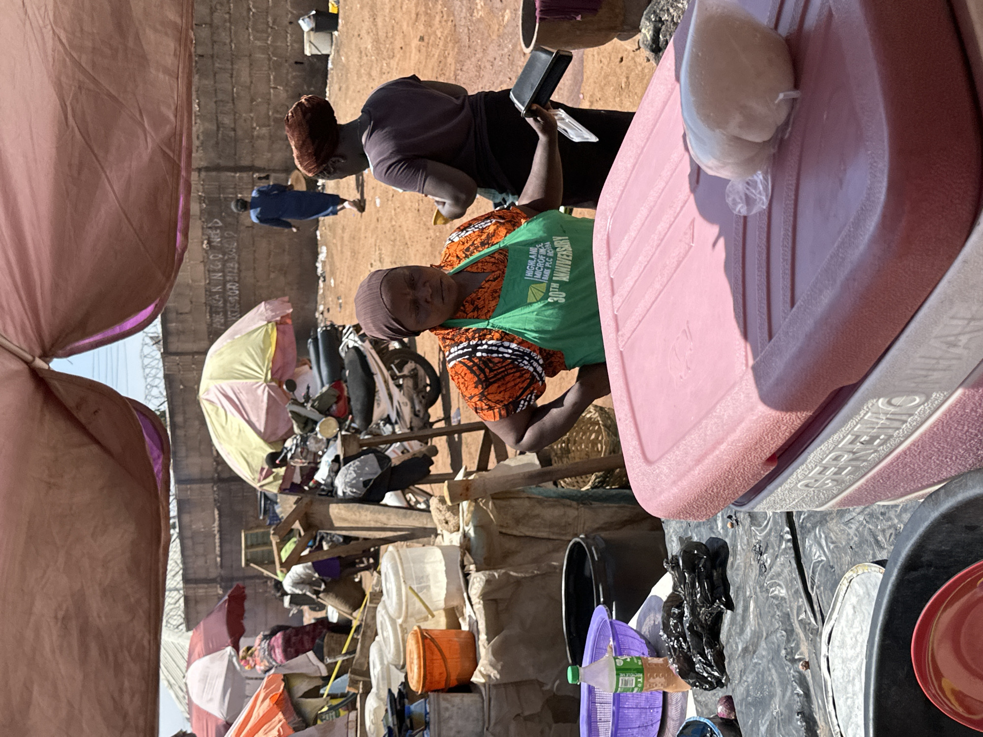 This media file captures an active outdoor market scene in Gura Top, Nigeria, under the bright sun of a clear day. The ground is dry and dusty, suggesting a dry season.
In the foreground, a woman is prominently featured, wearing an orange patterned top and a green apron. The apron clearly displays the text "HIGHLAND MICROFINANCE BANK PLC KADUNA 30TH ANNIVERSARY". She looks towards the viewer with a calm expression, positioned behind a large pink cooler that has the word "SUPERSAVER" embossed on its side. On top of this cooler, a small, white, plastic-wrapped item is visible. To her right, another woman in a dark top and head covering is partially visible from behind, seemingly engaged in a transaction or holding a small dark tray.
The immediate surroundings of these women include various market items such as plastic buckets, baskets, and other containers, indicating a stall selling diverse goods. In the middle ground and background, the market stretches further with numerous other vendors. Colorful umbrellas, primarily yellow-pink and red-white, provide shade over additional makeshift stalls constructed from wood and fabric. People can be seen moving around, suggesting a bustling commercial atmosphere. A distant concrete wall bears visible graffiti, including the partial text "ARE KAN NGO NEED ICES FOR 2023" followed by some numbers like "3004 3600". The bright, direct sunlight indicates a time of day around late morning or early afternoon.
