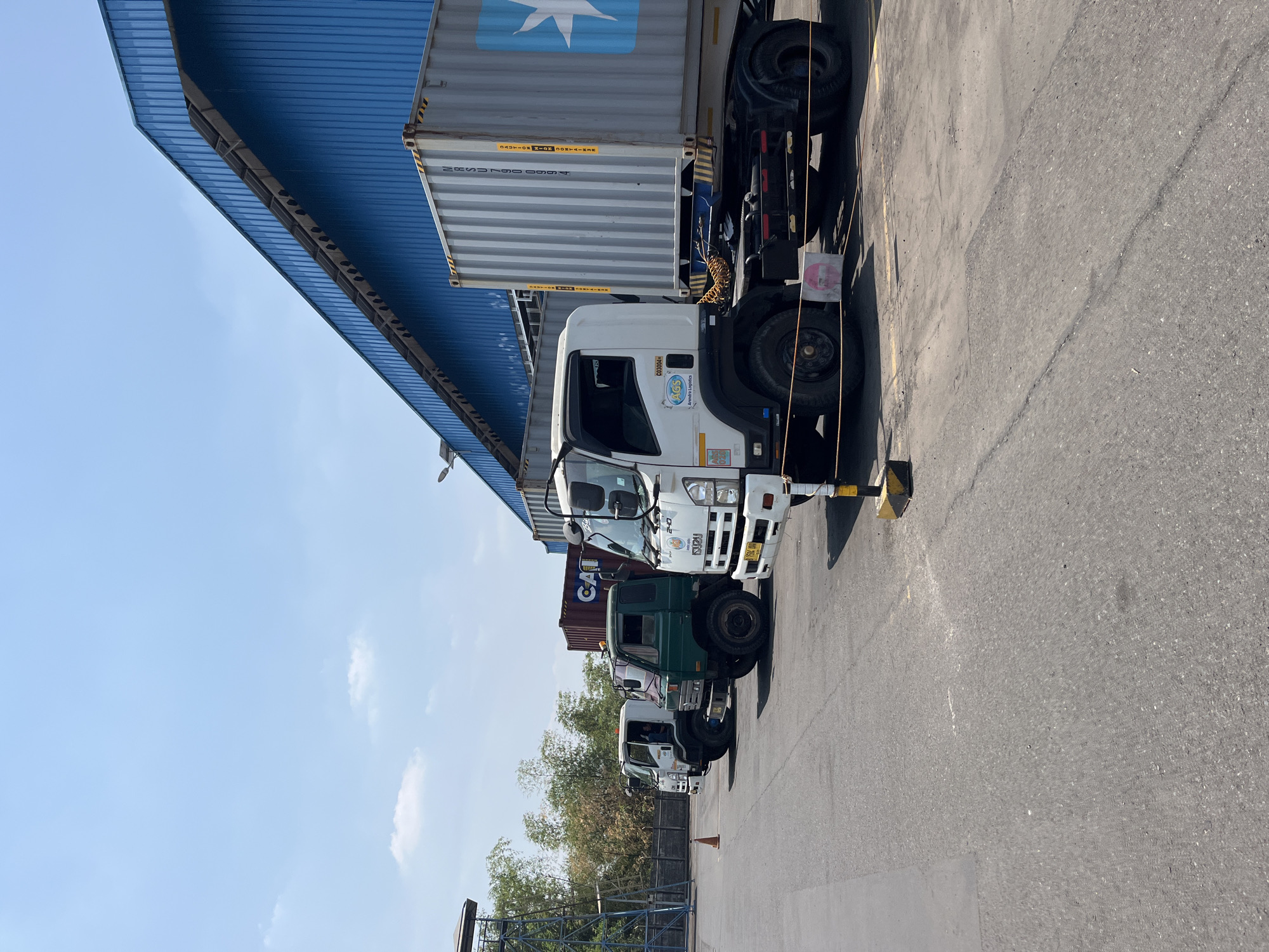 The image depicts a line of trucks parked in an outdoor lot, likely at a logistics or industrial facility in Pelang, Indonesia. The trucks are of various types, including a white Isuzu truck with a container, a green truck with a driver visible inside the cab, and another white truck. The environment includes a large, blue-sided building with a sloped roof, a clear blue sky with some clouds, and a paved lot with some tire marks and markings. There are also some trees and a fence visible in the background. One truck has a large shipping container with the Maersk logo. The scene is well-lit, suggesting it was taken during the day.