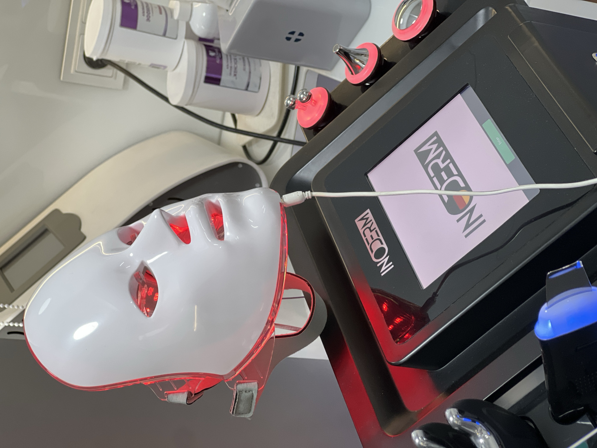 The image depicts a beauty treatment device, possibly in a spa or salon setting. The device features a white face mask with red LED lights illuminating the eye and mouth areas, connected by a white cable to a black control panel. The control panel displays the word "INODERM." Additional attachments with pink accents are visible above the screen. The background includes white product containers, possibly related to the treatment. No people are visible in the frame. The city of Yerevan, Armenia cannot be confirmed from the image alone.