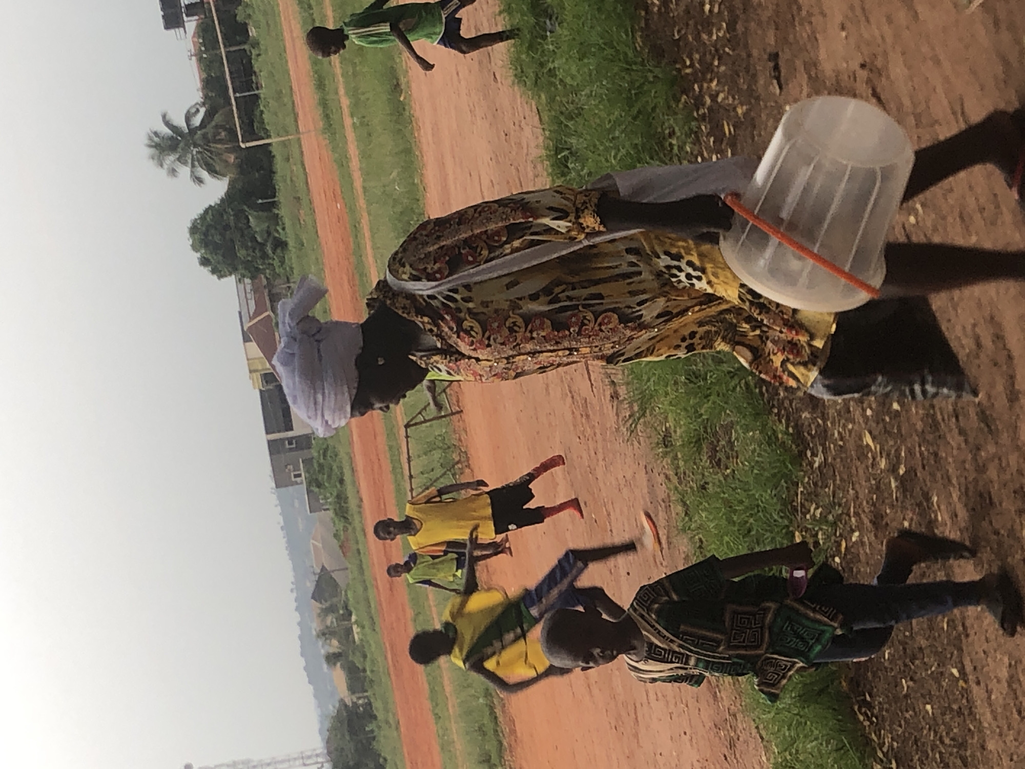 On a dirt field in Ukwu-Abwa, Nigeria, several people are present. An adult wearing a white head covering and a colorful patterned top carries a clear plastic bucket with an orange handle. Four children, wearing various brightly colored athletic shirts and shorts, are scattered across the field, some running. The field is surrounded by green grass, with trees, buildings, and a transmission tower in the background.