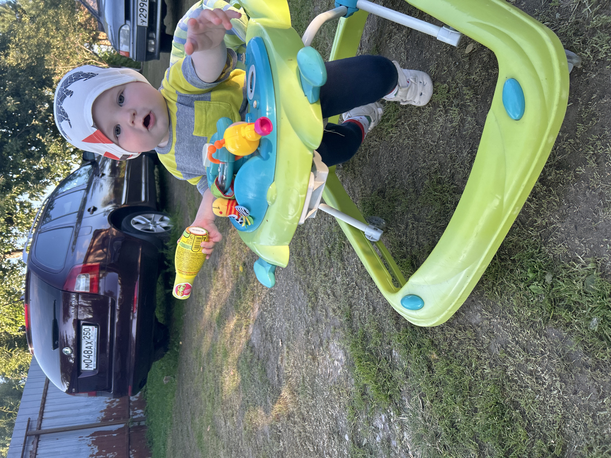 A baby is sitting in a green baby walker on a grassy area, holding a yellow bottle of drink. The baby is wearing a yellow and grey striped shirt and a white cap with a black and red pattern. In the background, a dark red station wagon car with a license plate reading "M048AX 250 RUS" is parked near a metal fence. To the right of the station wagon, a darker colored car is partially visible. The scene appears to be outdoors in a residential area.