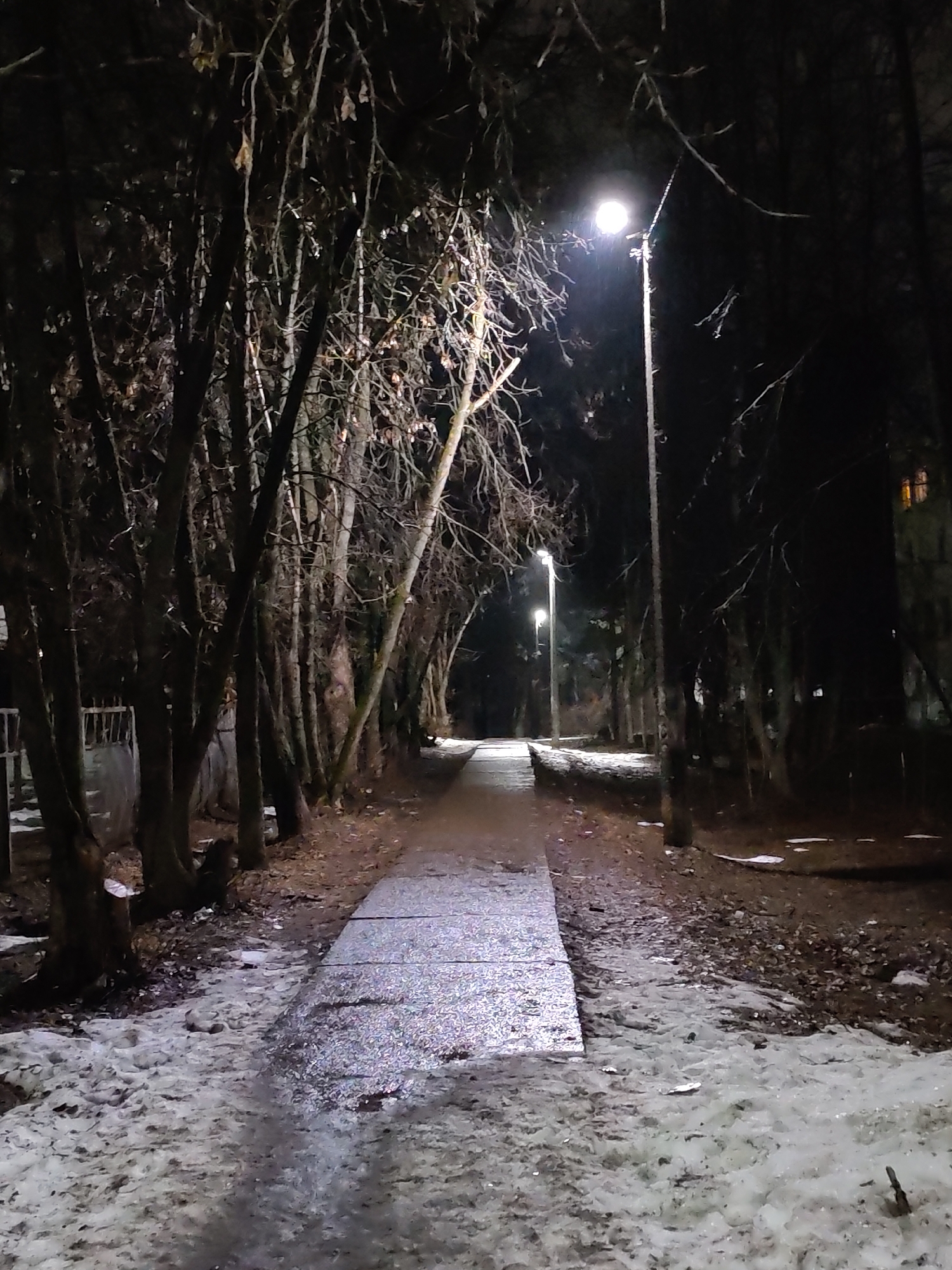 The image shows a dimly lit pathway in what appears to be a park at night. The pathway is composed of rectangular concrete slabs, and the ground beside it is partially covered with snow and leaf litter. Tall, bare trees line the pathway, their branches reaching into the darkness above. Streetlights illuminate the scene, casting a bright glow on the path and surrounding area, while leaving the edges of the image in shadow. A building with a lit window is partially visible on the right side of the frame. The image is likely taken in Chaikovsky, Russia, judging by the typical urban park environment and street lighting style. No people are visible in the image.