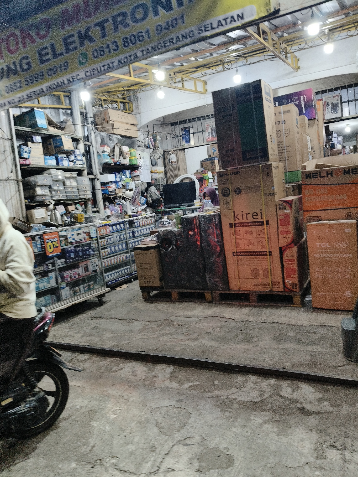 The image shows the exterior of a small electronics store in Tangerang Selatan, Indonesia. A sign above the entrance reads "TOKO MULA ELEKTRONIK" followed by phone numbers and an address that includes "CIPUTAT KOTA TANGERANG SELATAN." Inside the store, various electronic appliances are displayed on shelves and stacked on pallets. A person on a motorcycle is partially visible in the foreground, parked outside the store. The environment appears to be an urban street with a concrete floor.