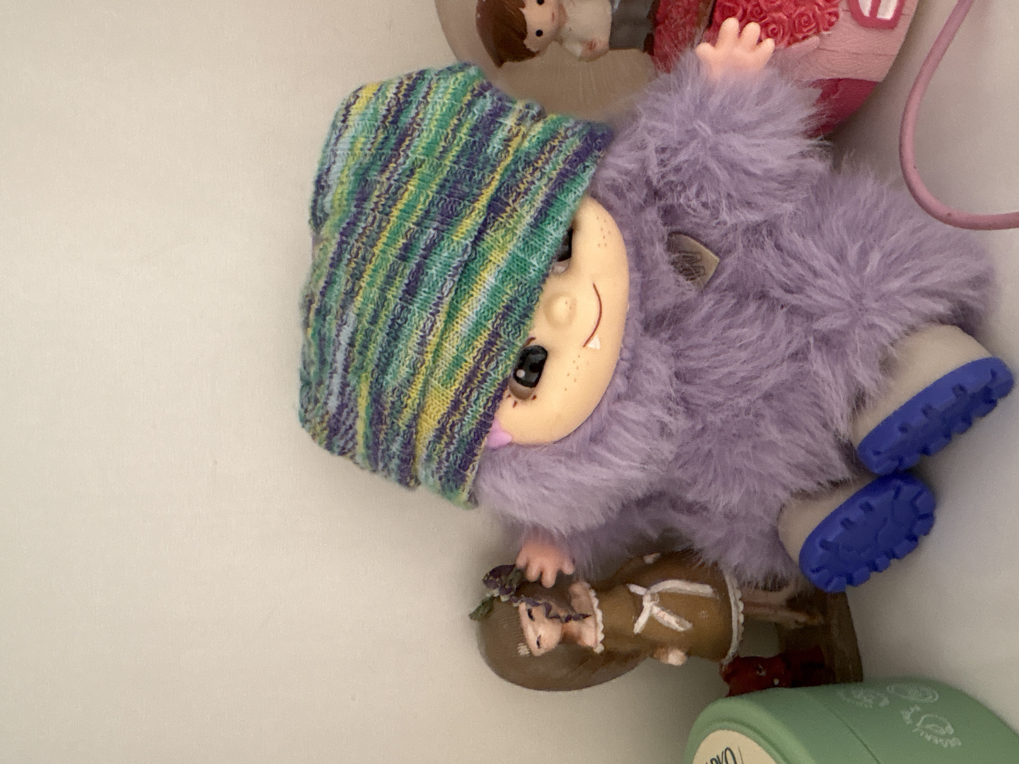 This is an indoor, eye-level, medium shot of a plush toy with purple fur and a knitted hat pulled down over its eyes. The toy is sitting and appears to be wearing blue shoes. To the left of the toy is a small, brown-haired doll wearing a tan dress. Further to the left and partially visible is a light green round container with a lid. To the right of the plush toy, a small figurine of a boy in a light-colored shirt and gray pants is visible. The background is a plain, light-colored wall. The scene is on a white shelf. There are no discernible elements within the image that would indicate the city of Baku, Azerbaijan.