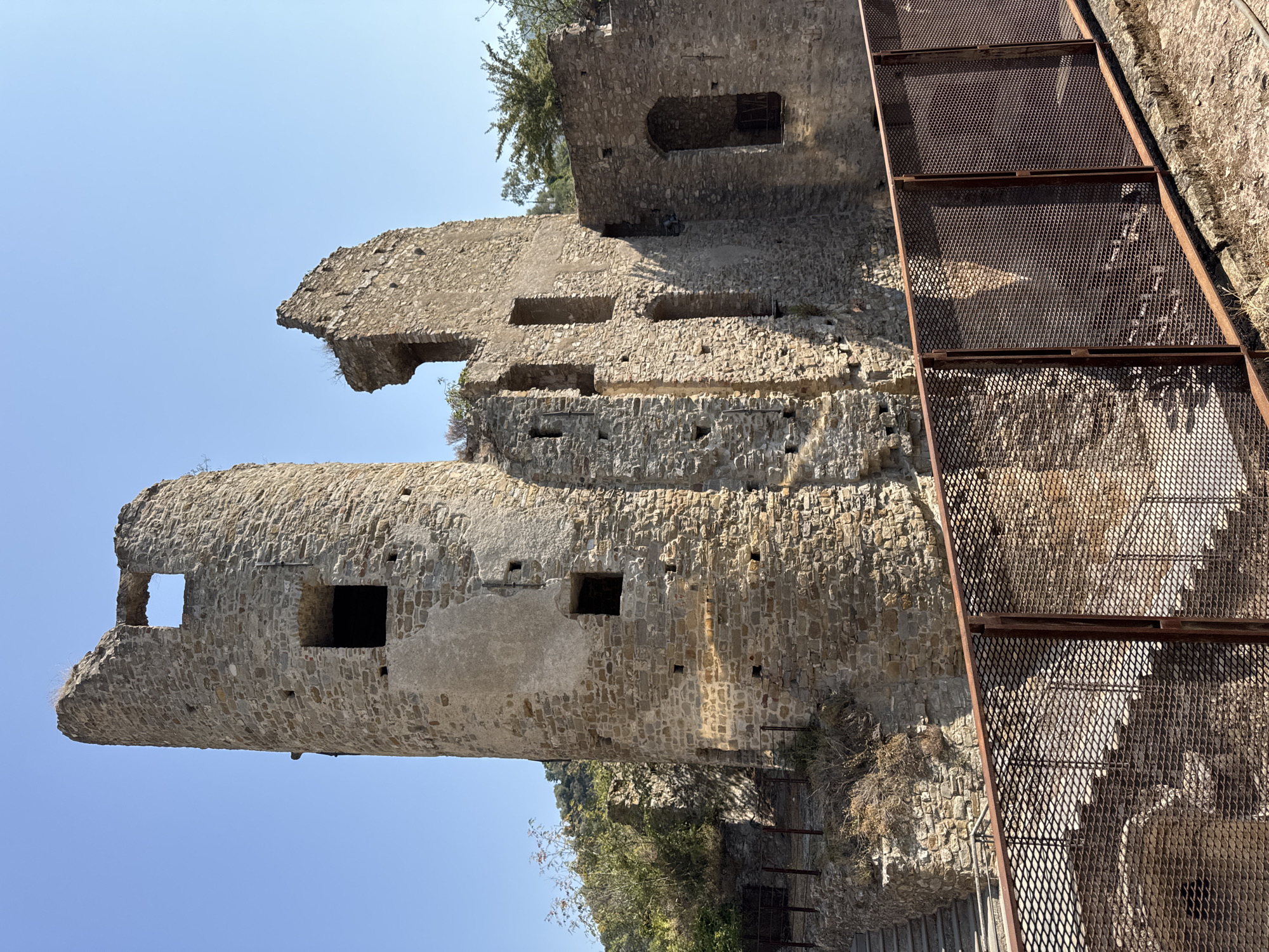 The image shows the ruins of a stone castle on a hillside under a clear blue sky. In the foreground, there is a rusted metal fence with a mesh pattern. Behind the fence, a stone staircase leads upwards towards the castle ruins. The castle itself is in disrepair, with crumbling walls and empty window openings. A cylindrical tower stands prominently on the left, its top section broken. Adjacent to the tower are sections of the castle's main structure, also showing significant decay. Vegetation, including trees and shrubs, grows around the ruins and on the hillside. The scene appears to be a historical site, likely open to visitors, as indicated by the presence of the fence and stairs. The location is the Doria Castle in Dolceacqua, Italy.