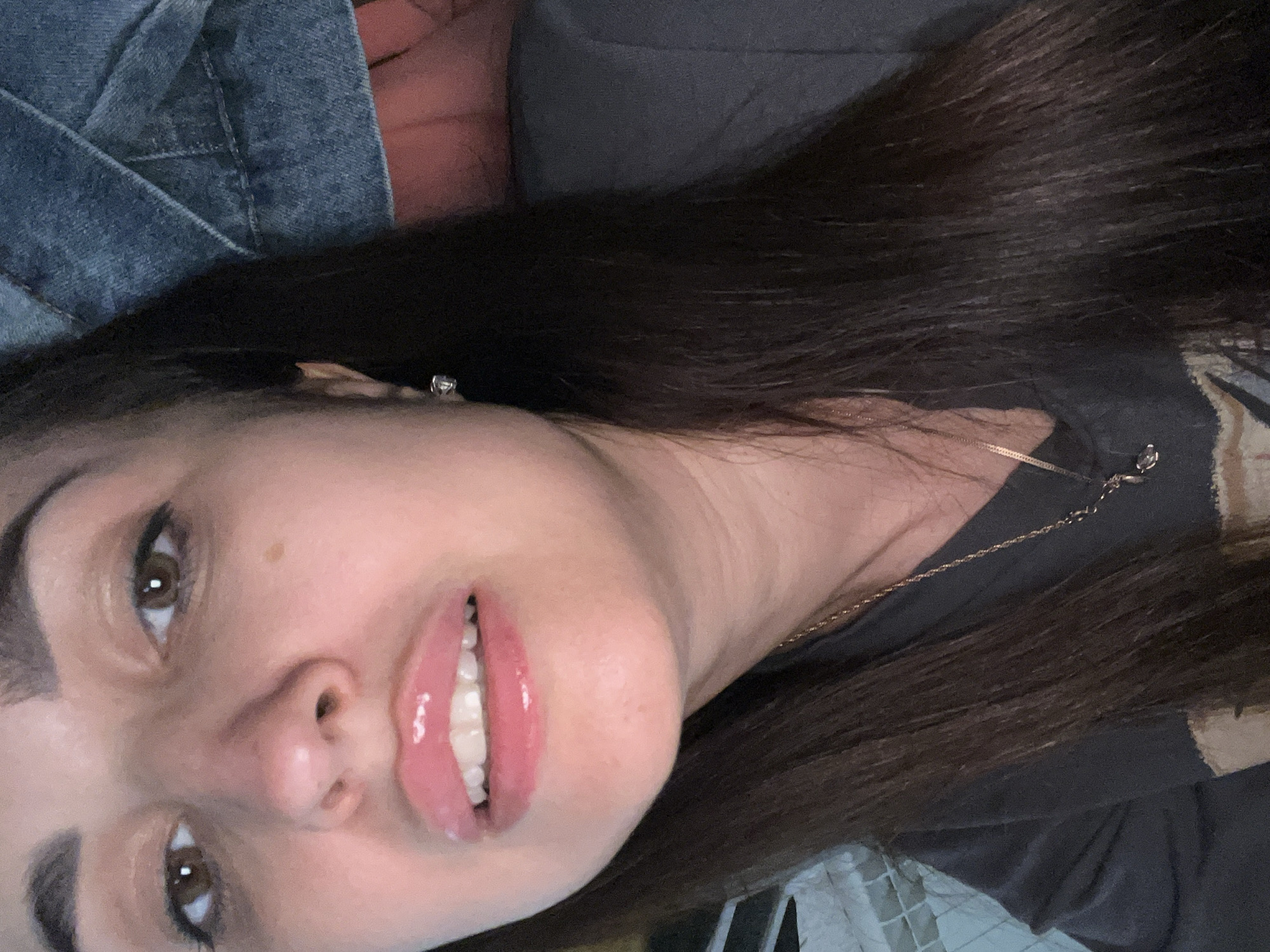 A close-up shot shows a woman's face, with her head tilted slightly. She has dark hair and brown eyes. Her lips are glossy and pink, and she is wearing a dark shirt with a gold necklace. The background is blurred, with hints of denim and fabric.