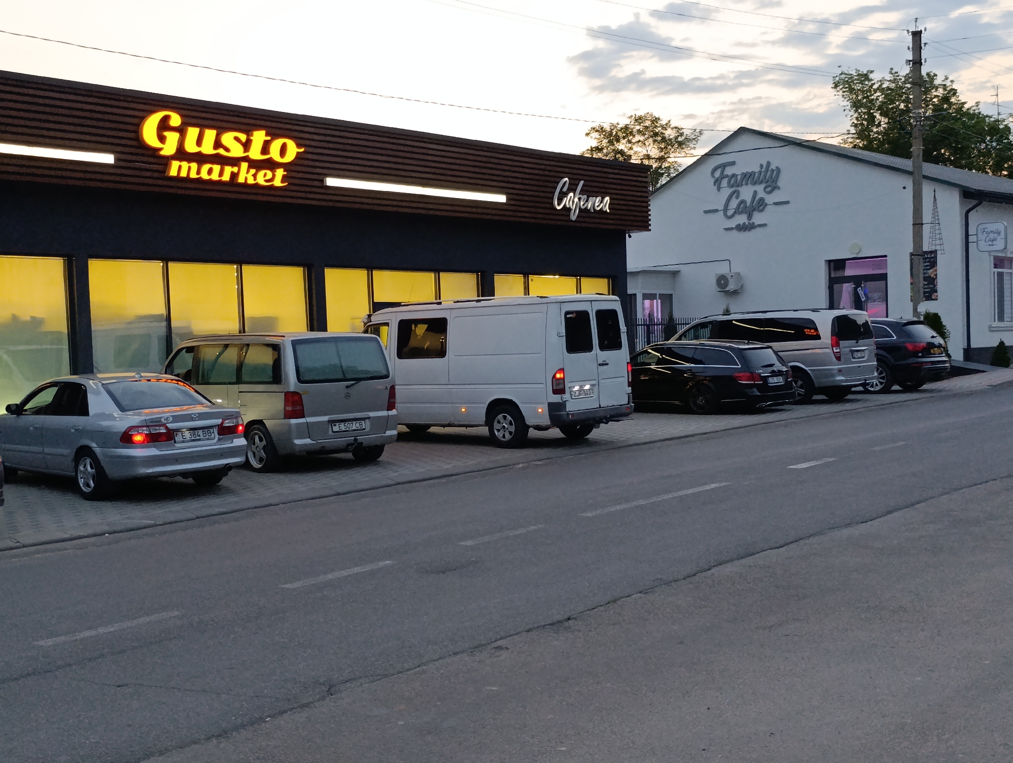 The image depicts a street scene in Cocieri, Moldova, with several vehicles parked in front of commercial establishments. On the left, a building with a dark facade and a prominent "Gusto market" sign in yellow lettering is visible. To its right, another building features a sign reading "Cafenea" in a stylized font. Further down the street, a white building displays "Family Cafe" in grey lettering. Parked vehicles include a silver sedan with a license plate beginning "E 384 BB", a silver minivan, a white cargo van, and two dark-colored SUVs. The environment appears to be dusk or early evening, with artificial lighting from the establishments and streetlights illuminating the scene. The sky is cloudy, suggesting an overcast condition. The road surface is asphalt, with visible lane markings.