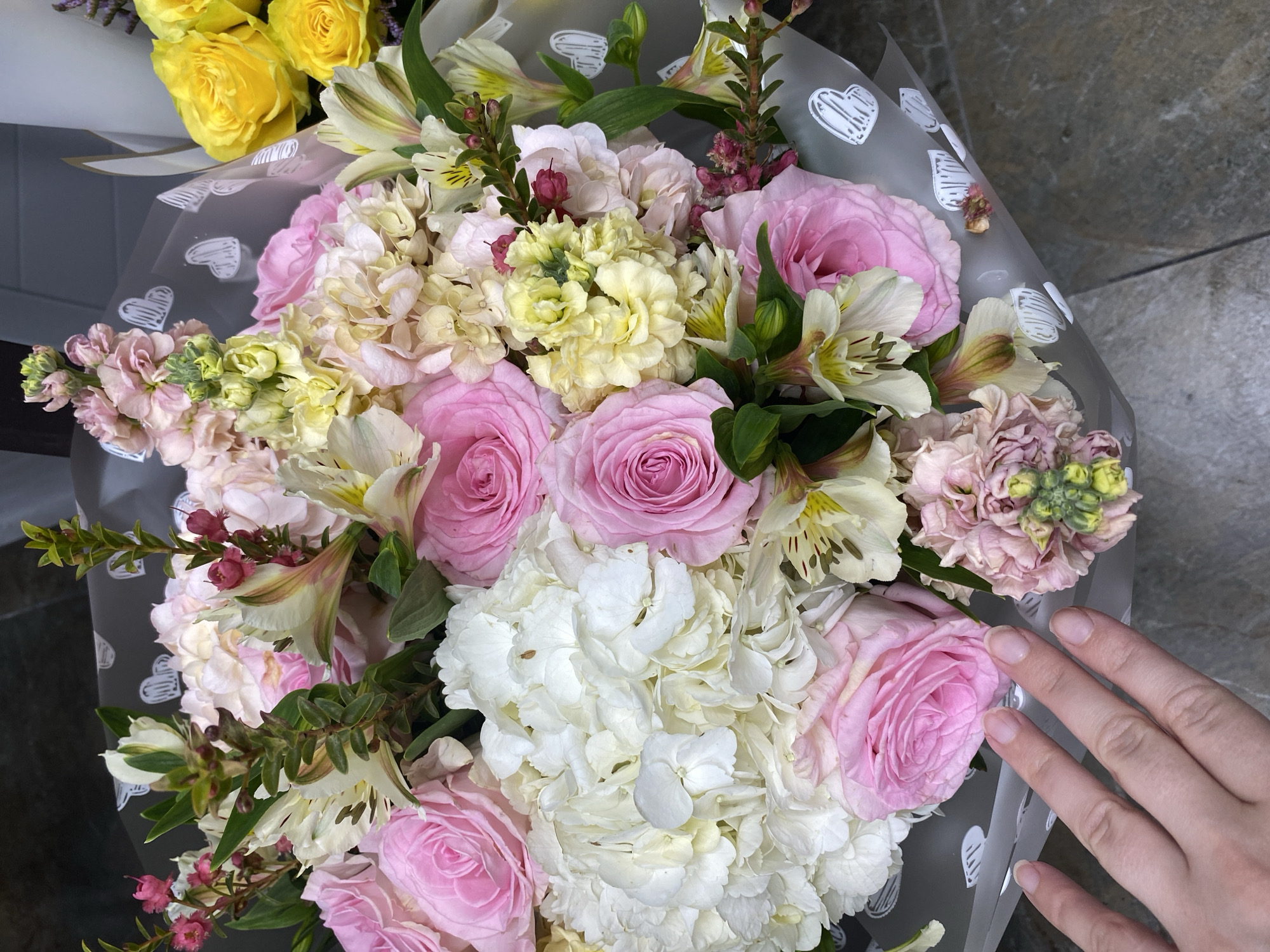 A bouquet of flowers is being held in someone's hand. The bouquet contains pink roses, white hydrangeas, yellow flowers, and other flowers in various shades of pink and cream. The bouquet is wrapped in a clear plastic wrap with a white heart pattern. The background is a gray textured surface. The location is not discernable from the image.