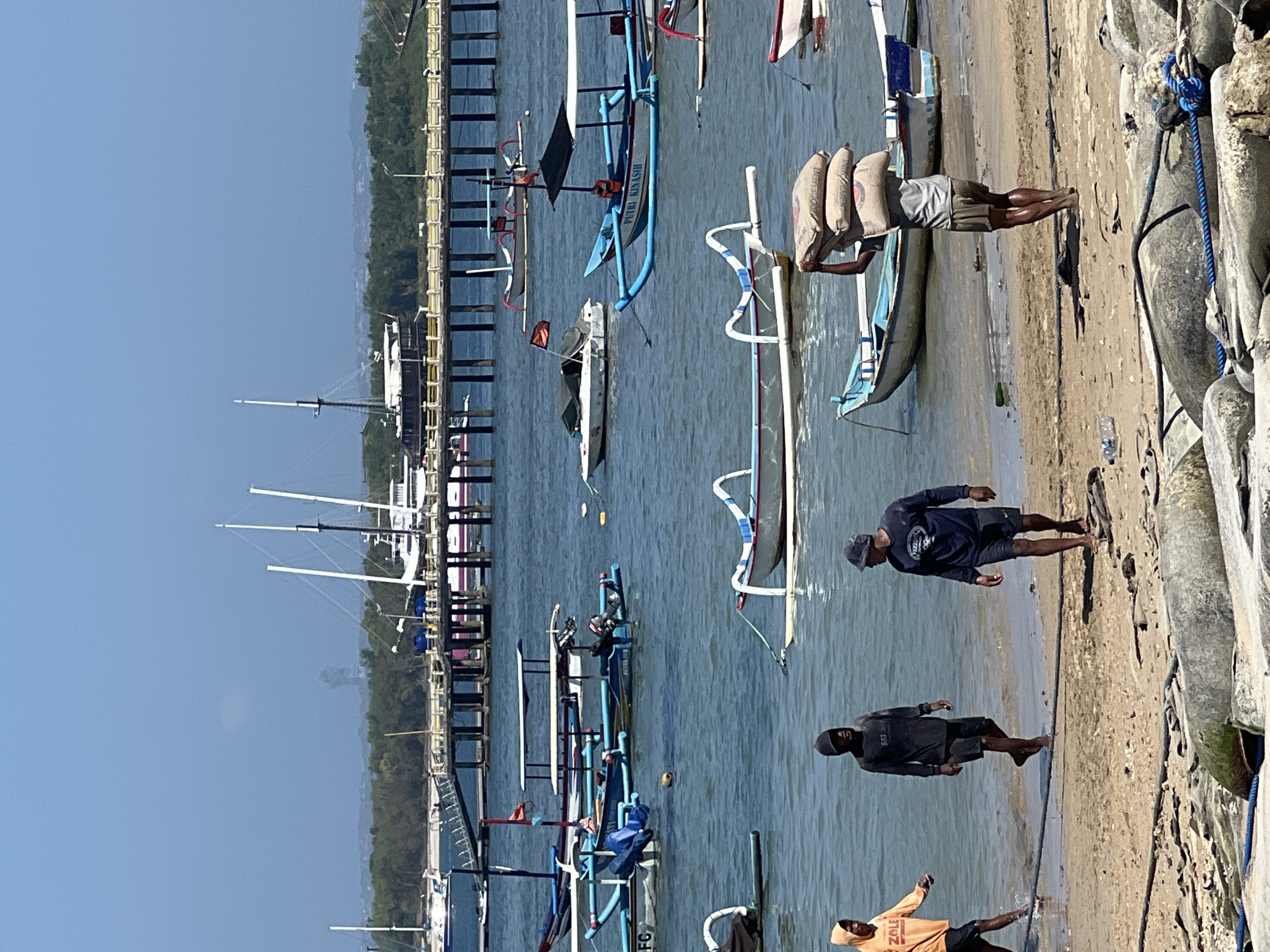 In Denpasar, Indonesia, this photograph captures a scene by the water on a sunny day. The foreground shows a sandy beach littered with rocks and ropes. Three men are walking on the beach; one in an orange hoodie with "ZOLE" on it, another in a black hooded shirt, and a third in a dark blue long-sleeved shirt with a logo. A fourth man in the middle ground is carrying two large sacks of what appears to be cement on his head, walking towards the right. In the water, numerous traditional Indonesian boats, known as outrigger boats, are anchored and moving. Some of these boats are blue and white, others white with red accents. Further out, a wooden pier extends into the water, lined with more boats, including larger yachts and what appears to be a ferry or cruise ship in the distance. The background consists of lush green trees and a clear blue sky.