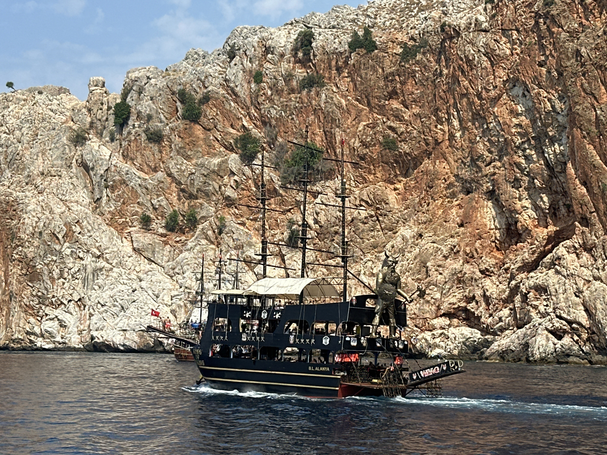 The image shows a black pirate-themed boat with multiple decks and masts sailing in the sea. The boat has a large statue of a horned figure on its bow and appears to be carrying passengers. In the background, a steep, rocky cliff face with sparse vegetation rises from the water. The sky is blue with some clouds. The text "BL ALANYA" is visible on the side of the boat, suggesting the location is Alanya, Turkey. The boat is moving to the right, leaving a wake in the water. Another smaller boat is visible behind the main boat. The overall scene depicts a scenic coastal view with a tourist boat.