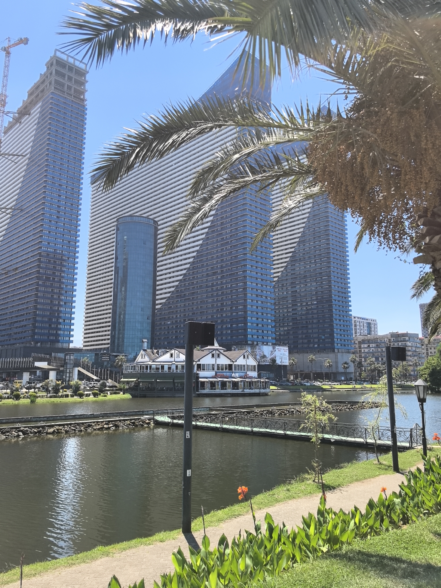 The image shows a waterfront scene in Batumi, Georgia, dominated by modern skyscrapers and a lake. In the foreground, a path bordered by lush green grass and flowering plants leads towards the lake. Orange flowers are visible among the foliage. A lamppost stands beside the path. The lake itself is dark and reflects the sky and surrounding buildings. A decorative metal bridge with a green railing spans a section of the lake. On the far side of the lake, a white building with a distinctive architectural style, possibly a restaurant or hotel, is situated. Behind this building, imposing skyscrapers rise, characterized by their blue and white facades. One particularly tall skyscraper is under construction, with a crane visible at the top. Palm trees are also present, with fronds obscuring parts of the view. The sky is clear and blue, indicating a sunny day. The scene appears to be a public park or promenade area in Batumi, likely designed for recreation and offering views of the city's modern architecture.