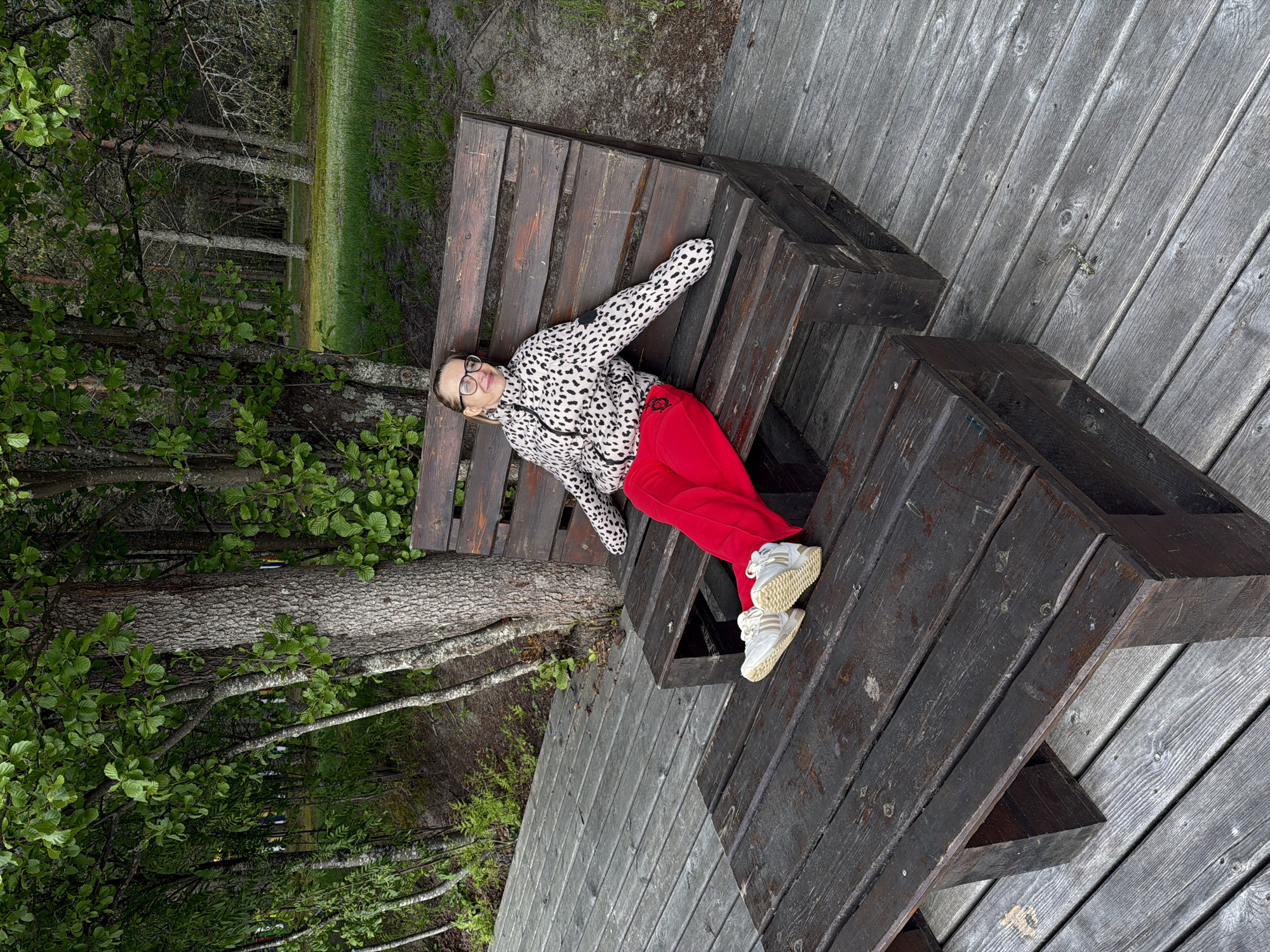 A person is lying on a dark wooden park bench. The individual is wearing glasses, a white and black spotted jacket, red pants, and white sneakers. The bench is placed on a light gray wooden deck. Trees and greenery are visible in the background. The location is Zaplav'e, Russia.
