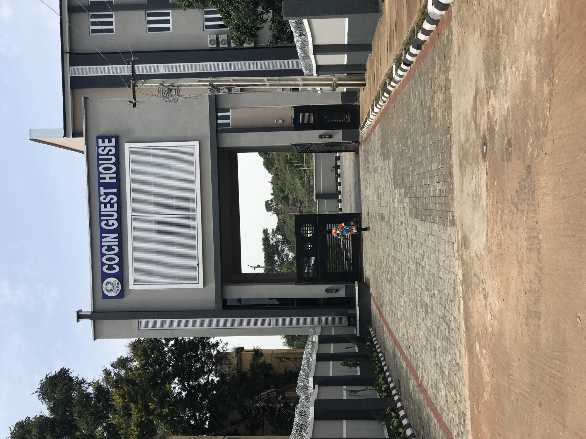 The image shows the entrance to "COCIN GUEST HOUSE" in Jos, Nigeria. The building is modern, with a grey facade and dark accents. A blue sign with white lettering identifies the establishment. Above the entrance, a large window or decorative panel with vertical white lines is visible. A person in a colorful patterned shirt is walking away from the camera towards the open gates of the guest house. The driveway is paved with interlocking bricks, bordered by a red line and black and white striped curbs. A barbed wire fence runs along the perimeter of the property. In the background, trees and a rocky hill are visible, characteristic of the landscape in Jos. The sky is overcast.