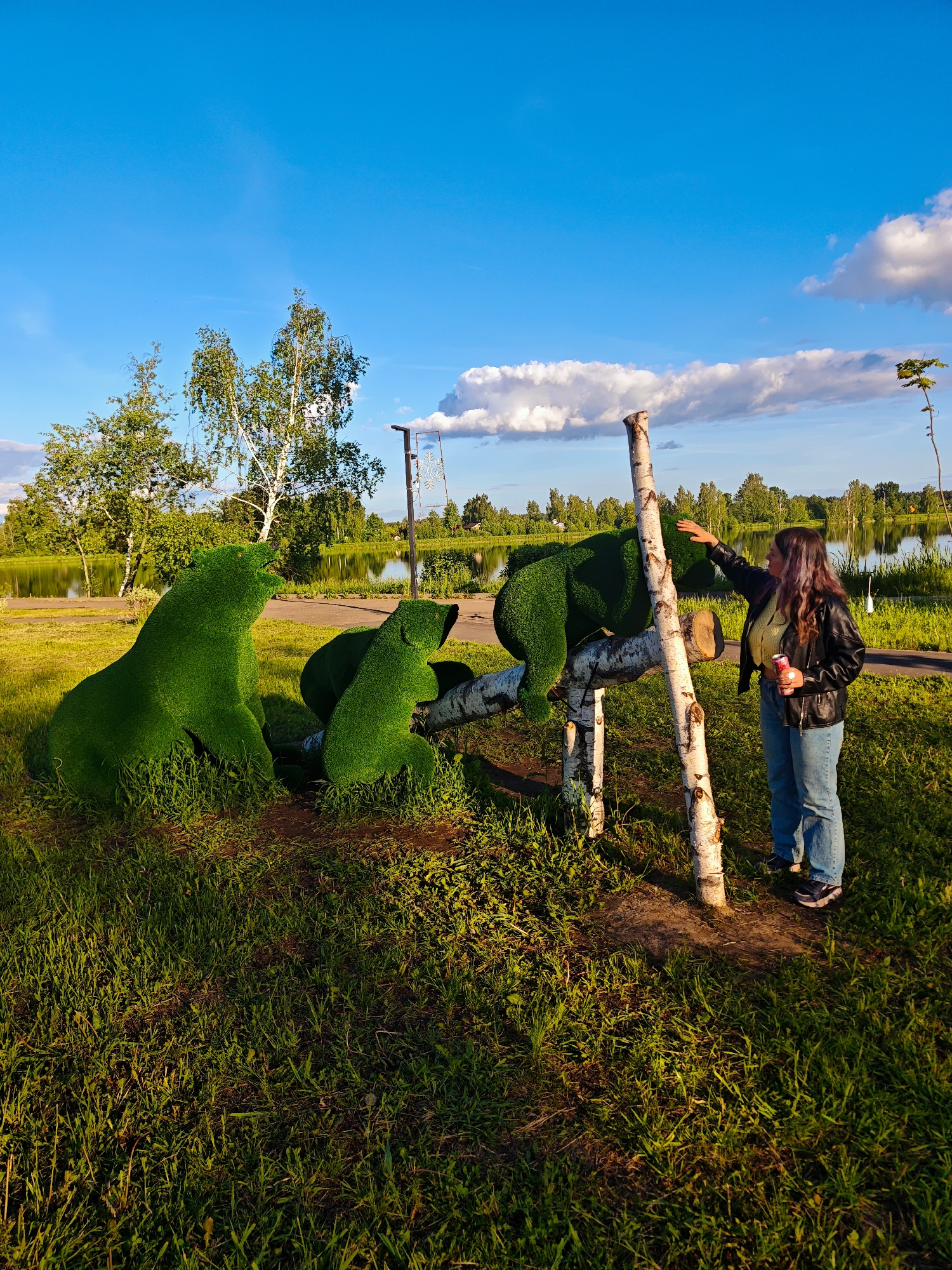 The image captures an outdoor park scene featuring a woman and topiary sculptures during the golden hour. A woman with long, wavy brown hair, dressed in a black leather jacket, light-colored top, blue jeans, and sneakers, is interacting with one of the topiaries. She gently touches the head of a large topiary bear with her right hand and holds a red "Coca-Cola" can in her left.
Three vibrant green topiary bears, sculpted from grass, are prominent. The largest bear is positioned on a rustic support structure made of three debarked white birch logs, one vertical and two horizontal. Two smaller topiary bears are seated on the lush green grass below, appearing to look towards the larger one.
The scene is set against a backdrop of a calm body of water, likely a lake or pond, which reflects the clear blue sky and the green trees lining its far bank. The foreground and immediate surroundings are covered in green grass, illuminated by the warm, low-angle sunlight of late afternoon or early evening, creating long shadows. A paved path is visible in the middle ground, running along the water's edge. In the background, distant trees and a street lamp with a decorative, possibly snowflake-shaped, element are visible. The sky is bright blue with scattered white clouds, indicating fair weather. The atmosphere appears peaceful and pleasant.